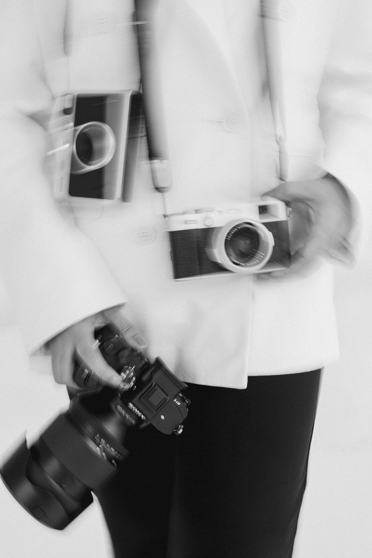 Black and white photo of a photographer holding multiple vintage and modern cameras while wearing a white blazer.