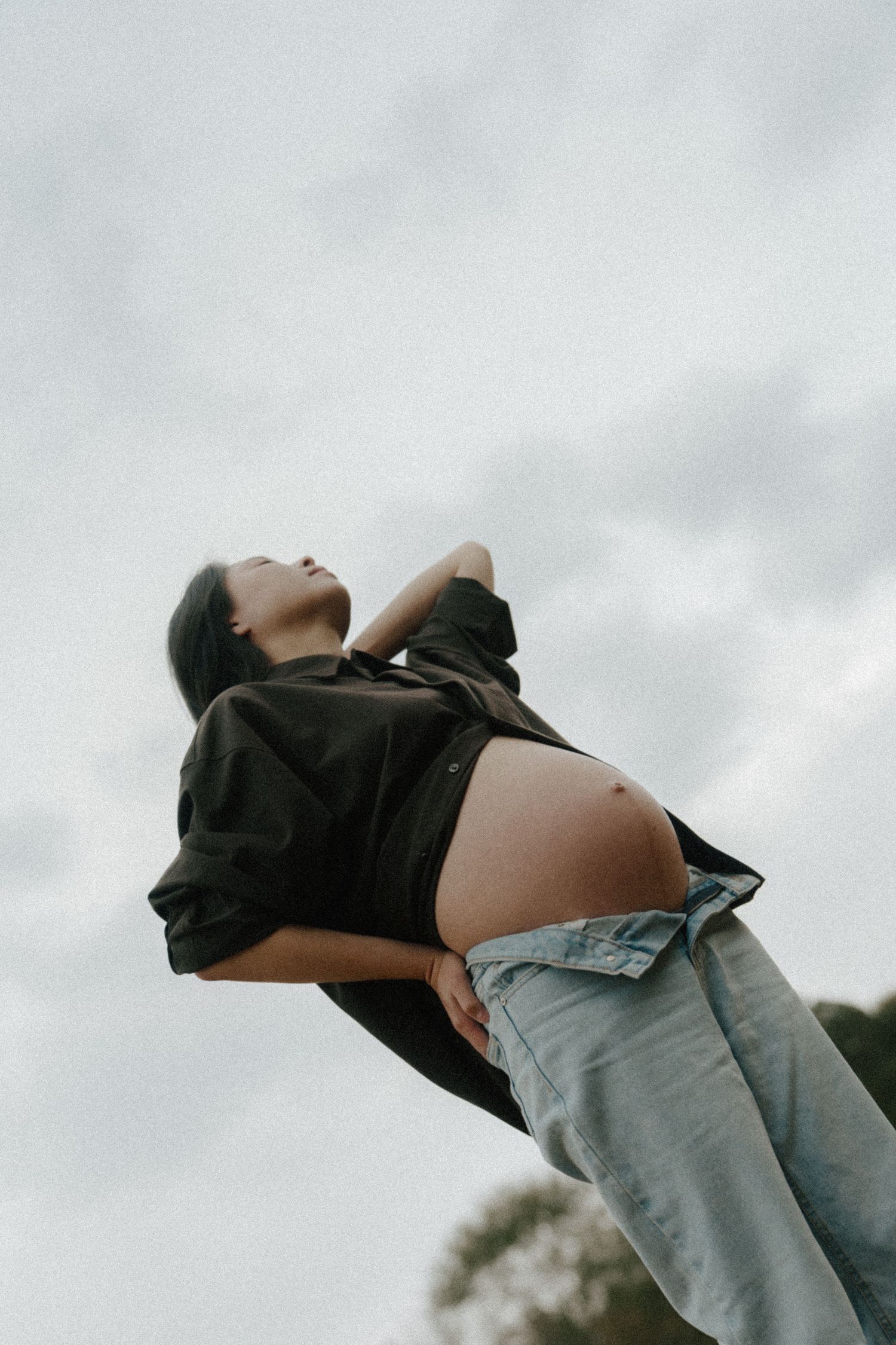 Pregnant woman in black shirt and jeans posing outdoors against cloudy sky, showcasing baby bump.