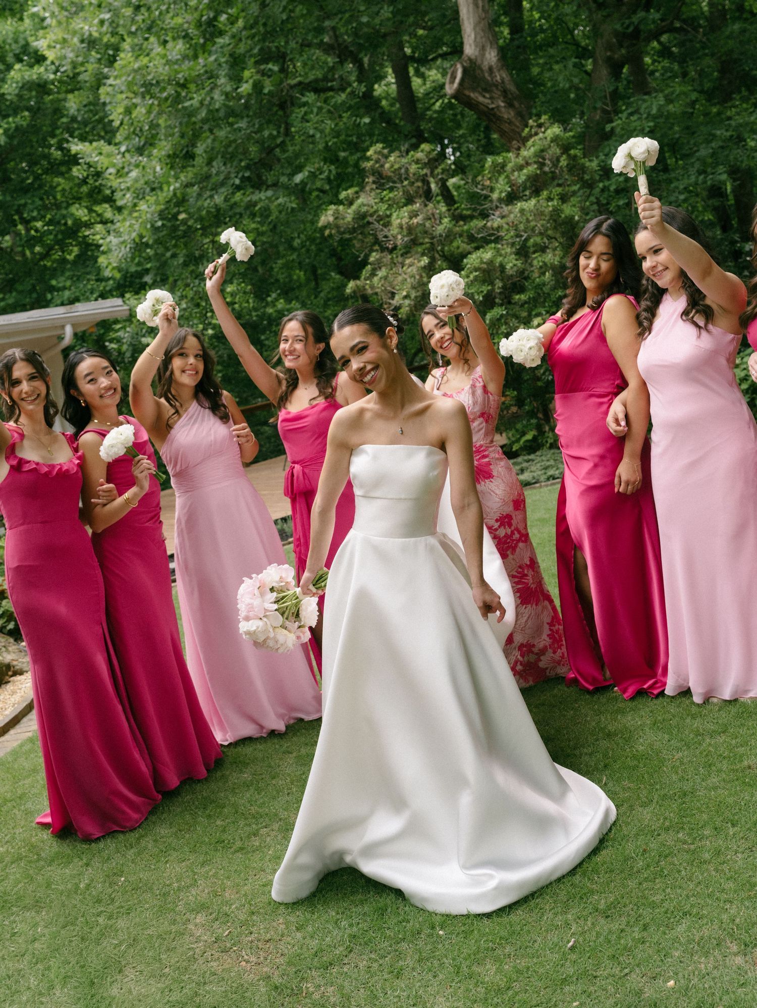 Bride in strapless white gown poses with bridesmaids in pink and fuchsia dresses holding white bouquets.