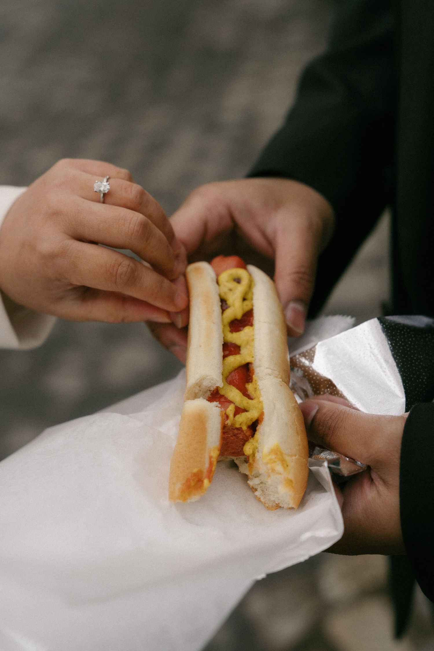 Newly engaged couple sharing a hot dog with mustard and ketchup, diamond engagement ring visible on woman's hand.