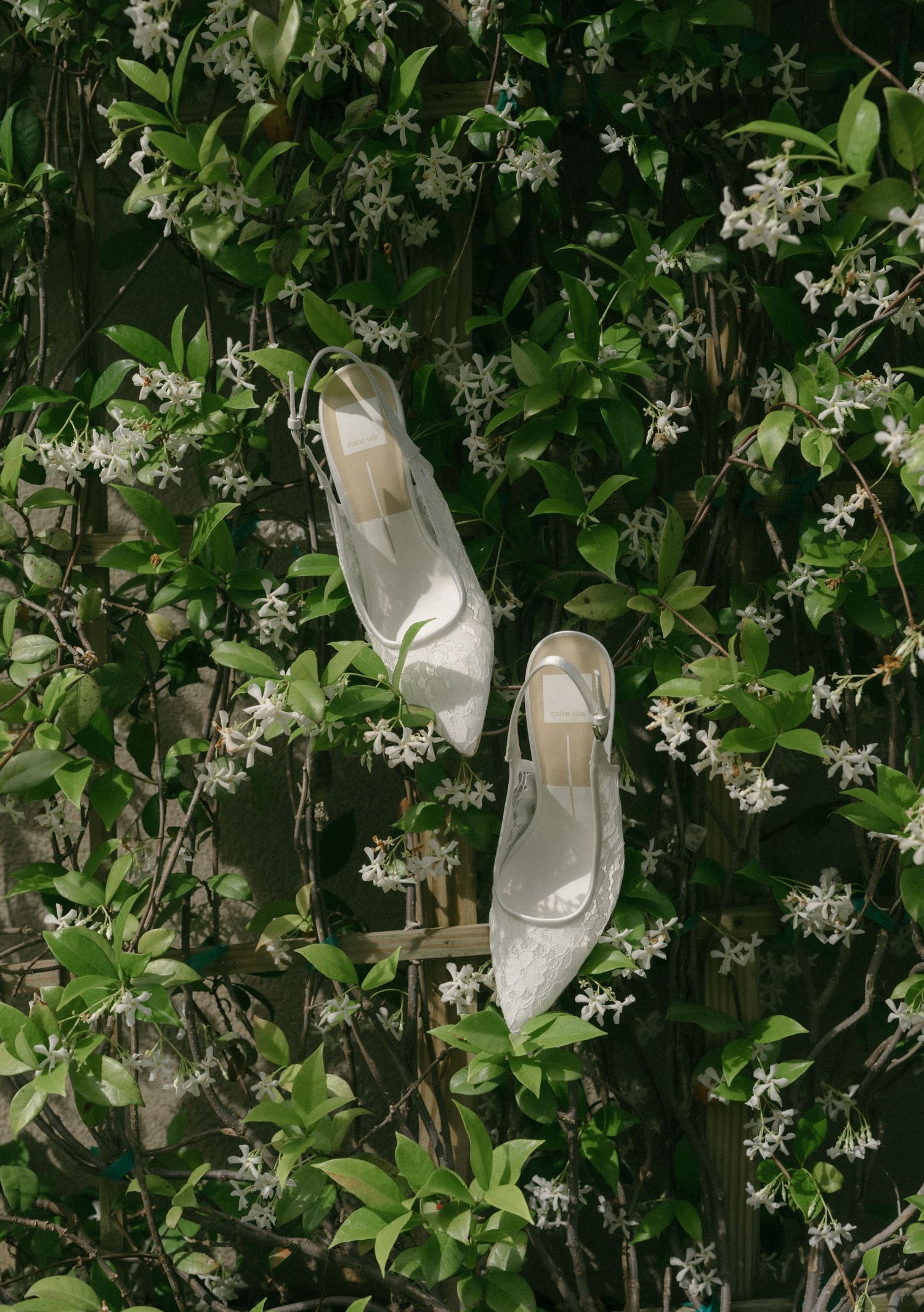White slingback heels resting among lush green foliage with delicate white flowers in a romantic outdoor setting.
