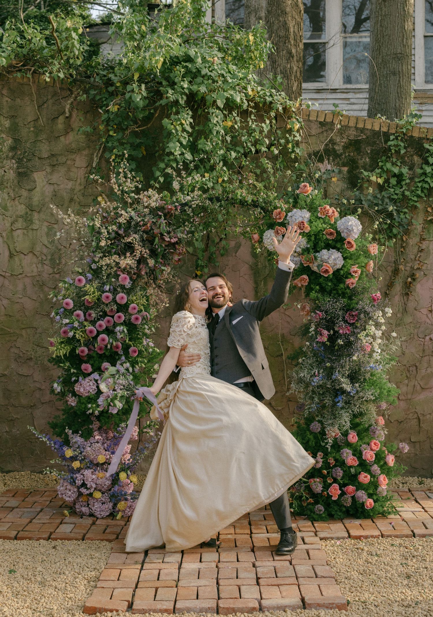 Couple celebrating under a lush floral arch with pink and white blooms at an outdoor wedding venue.