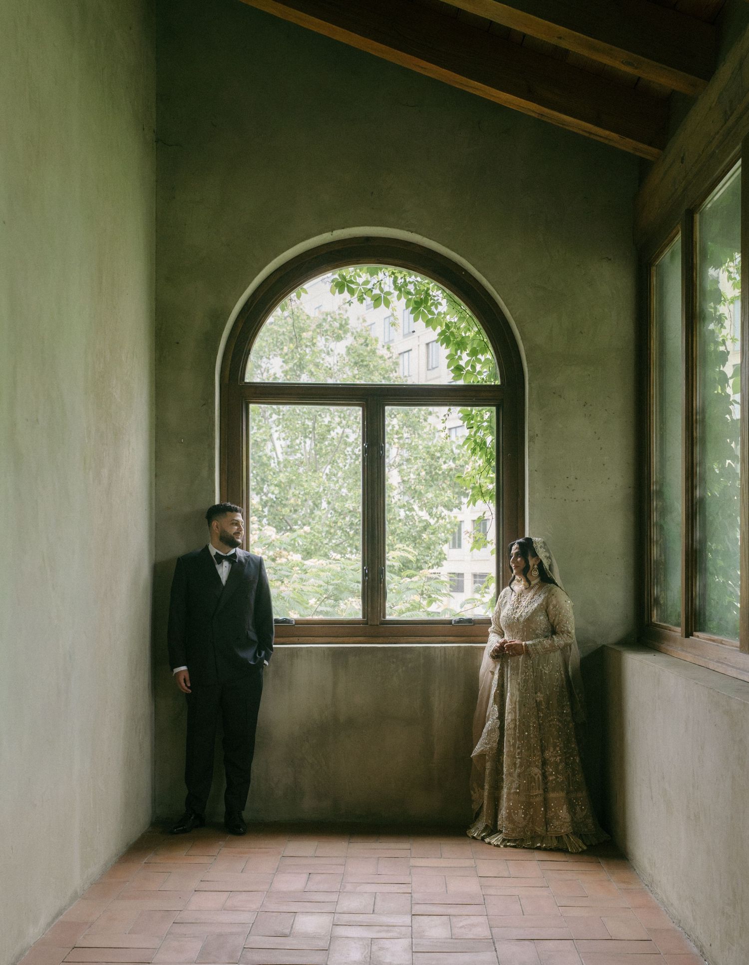 Couple in wedding attire standing apart in an arched green hallway with a large window and lush trees outside.