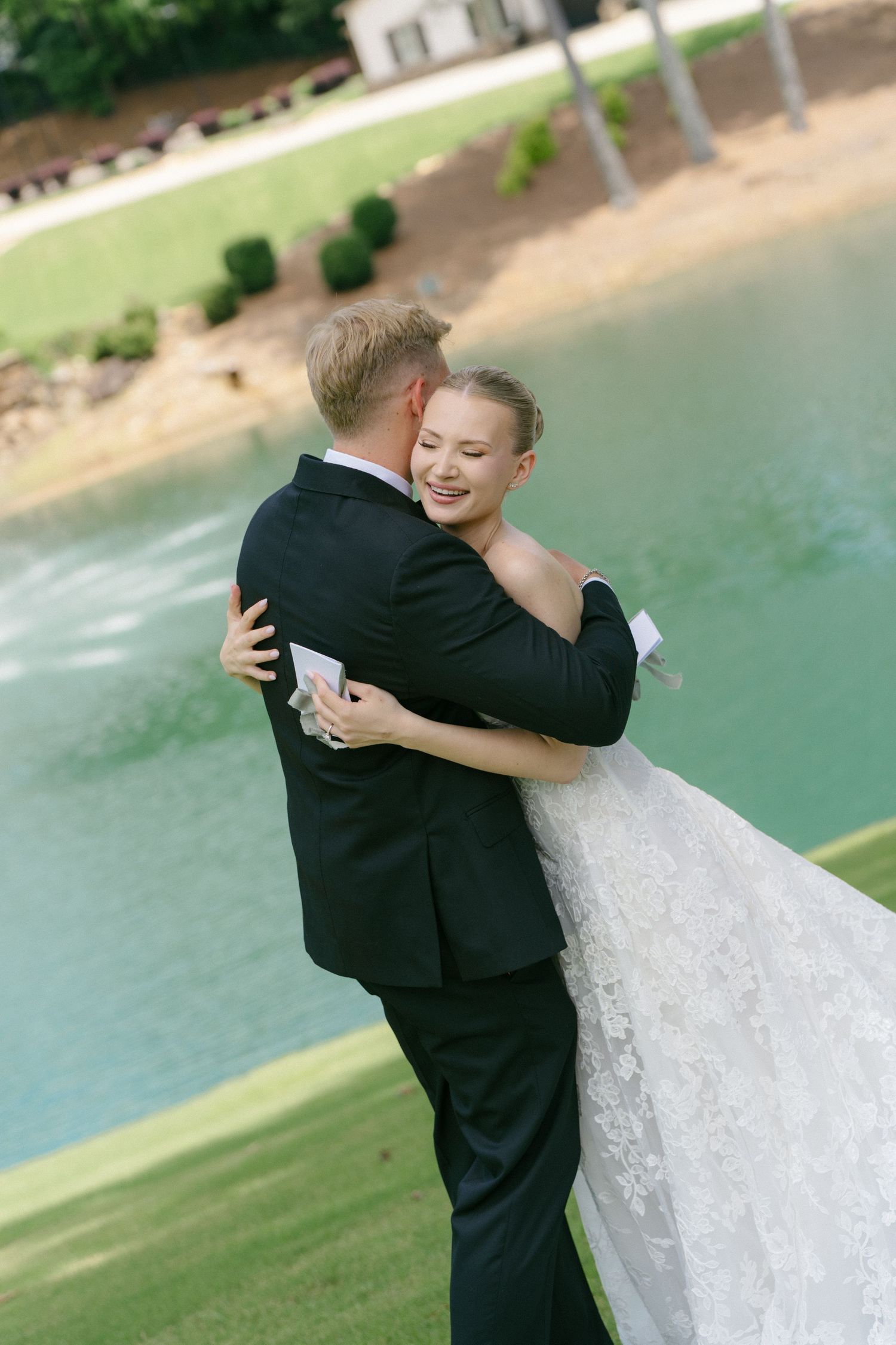 Bride and groom share a romantic embrace outdoors near a serene pond, bride smiling in a lace gown.