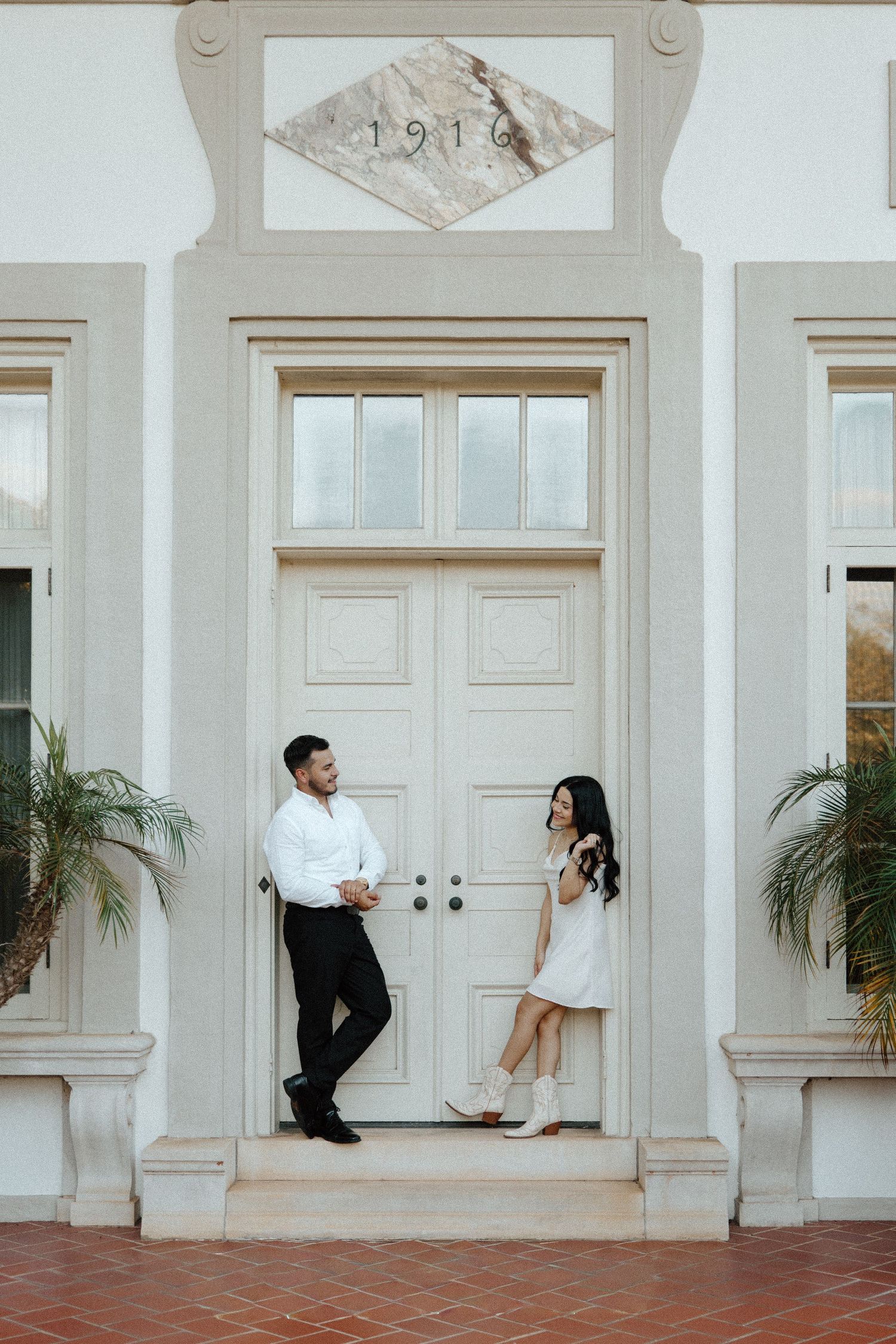 Couple in white outfits posing by a elegant cream doorway with 1916 above, sharing a romantic moment.