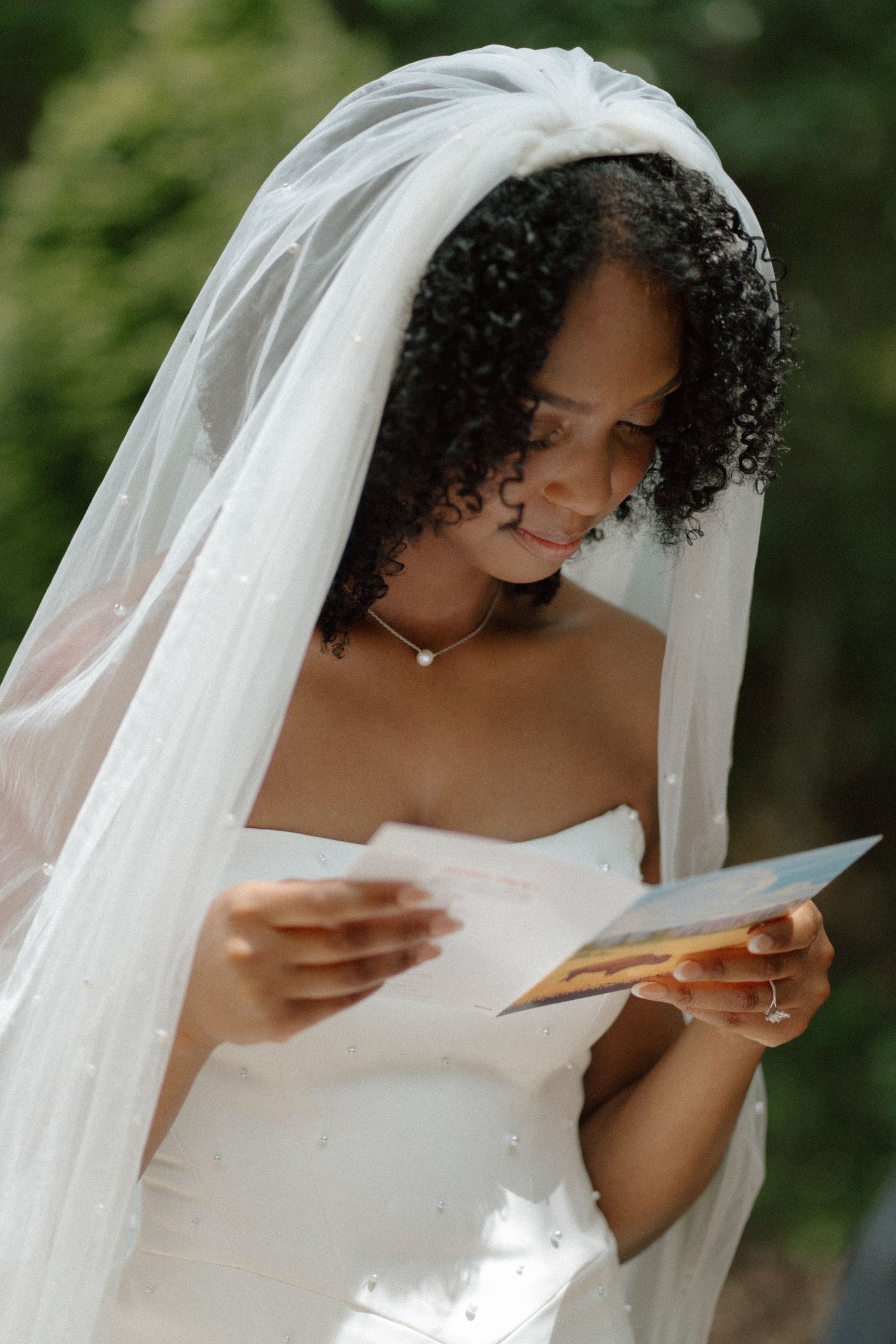 Beautiful Black bride in white strapless gown and veil reading a heartfelt letter outdoors.