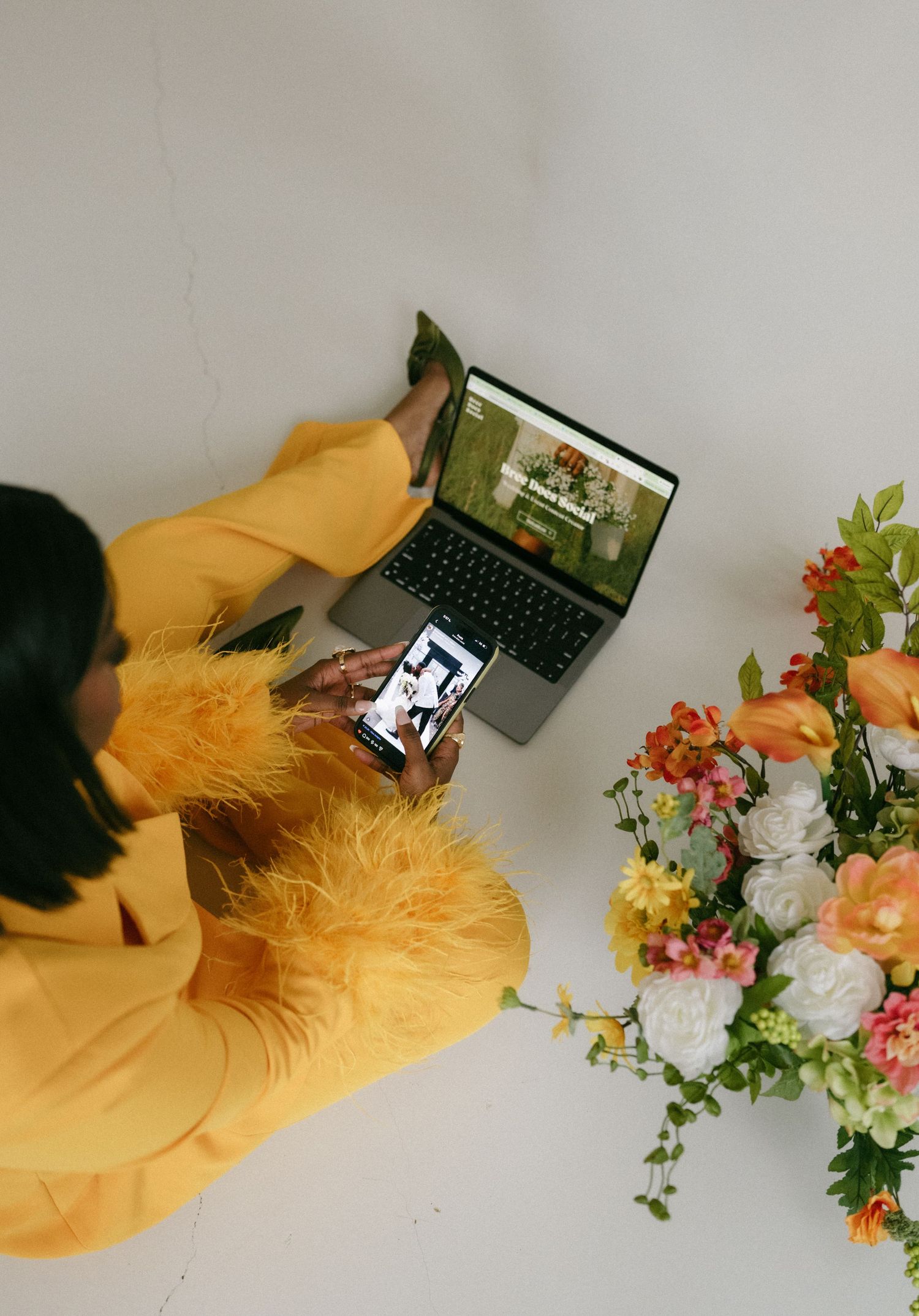 Woman in yellow outfit using smartphone and laptop beside colorful floral arrangement, viewed from above.
