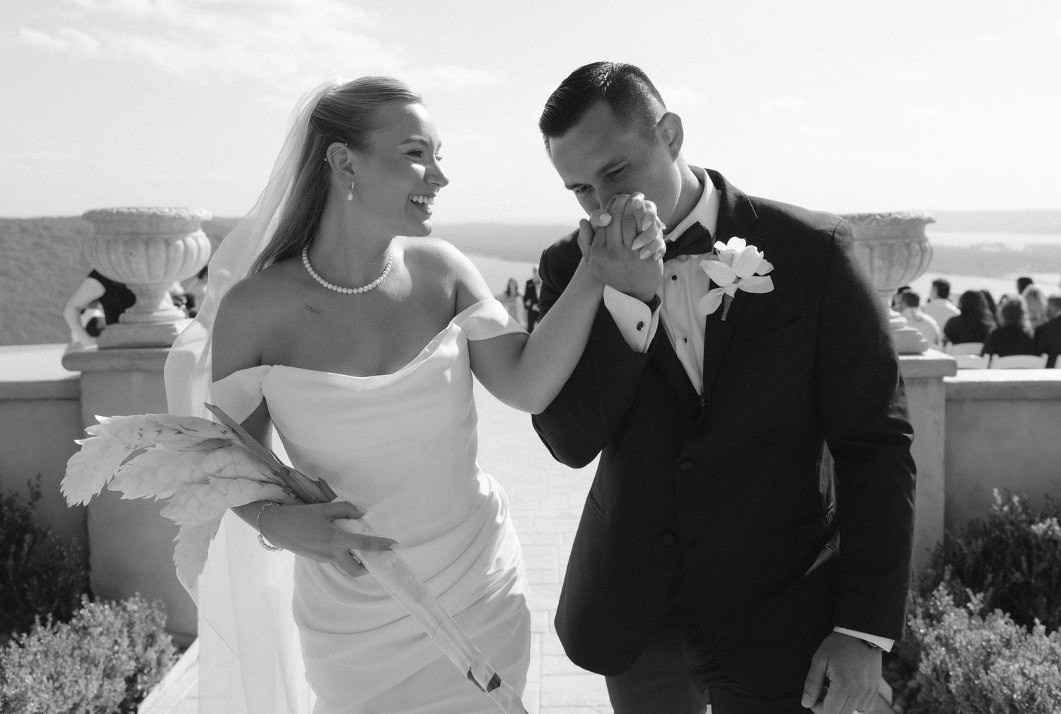 Black and white photo of groom kissing bride's hand at outdoor wedding ceremony with ocean view.