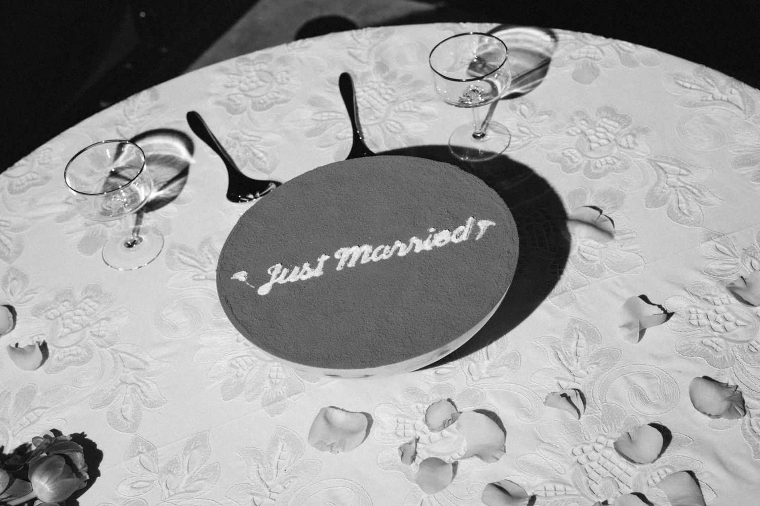 Black and white photo of a Just Married sign on a white tablecloth with rose petals and wine glasses.