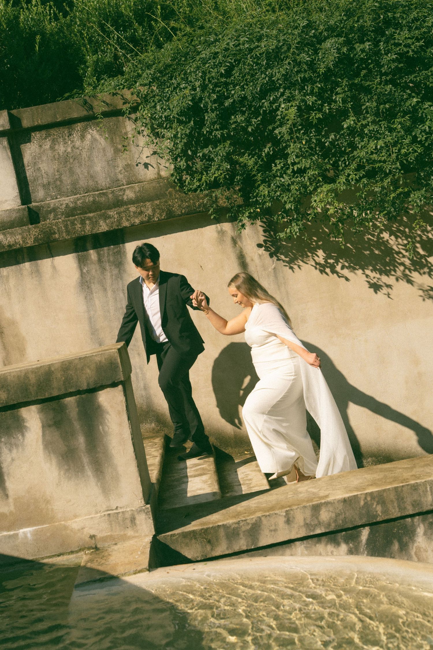 Bride in white dress and groom in black suit hold hands while climbing concrete steps near water.