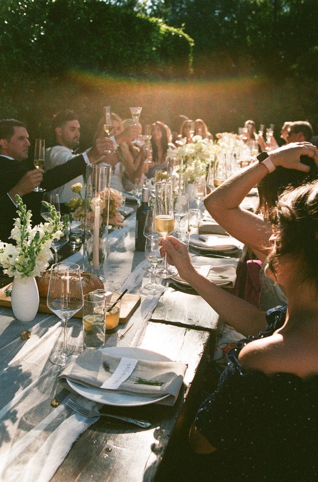 Guests raising glasses in a toast at an elegant outdoor long table dinner party with floral arrangements and golden sunlight.