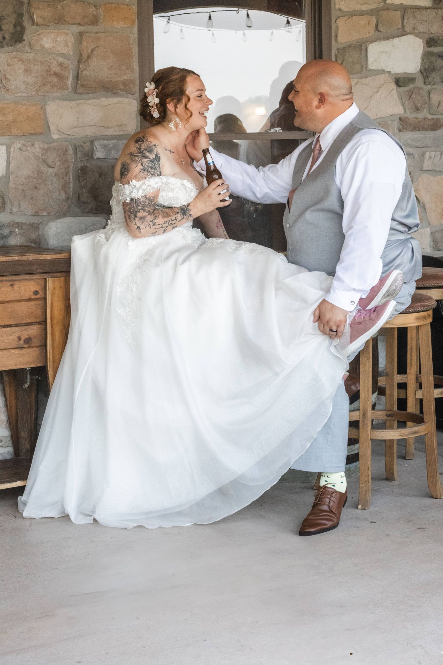 Bride in white gown and groom in gray vest share a joyful moment at a rustic stone venue wedding.