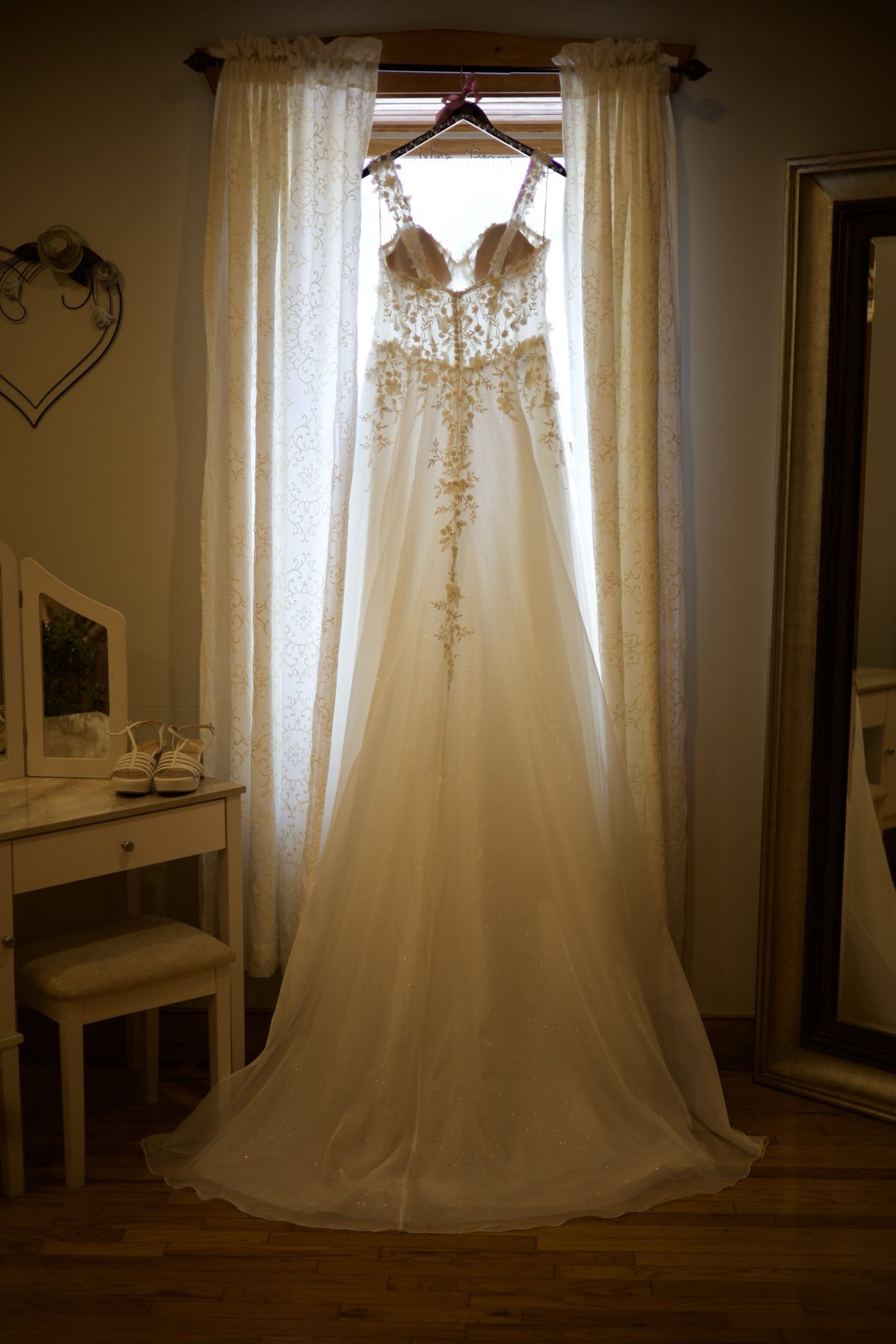 Elegant lace wedding dress hanging in window with white curtains and vanity table nearby.