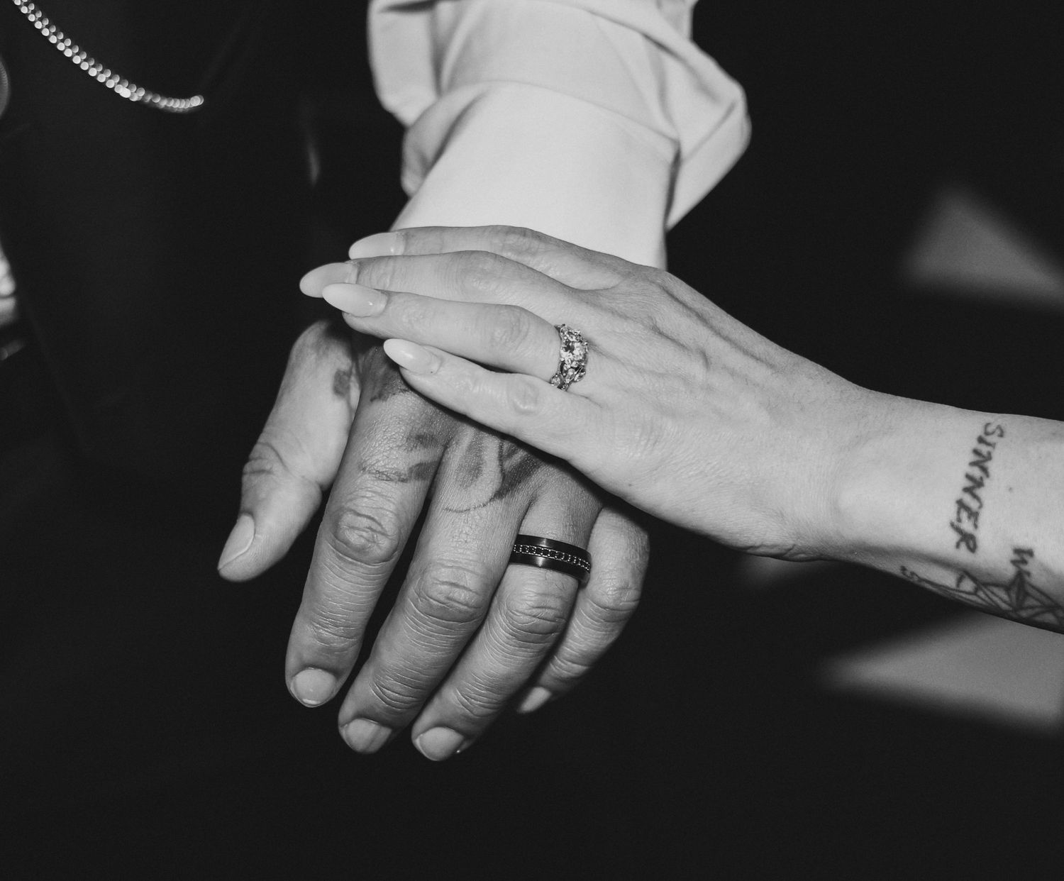Black and white photo of three hands stacked together, showing wedding rings and tattoos, symbolizing love and unity.