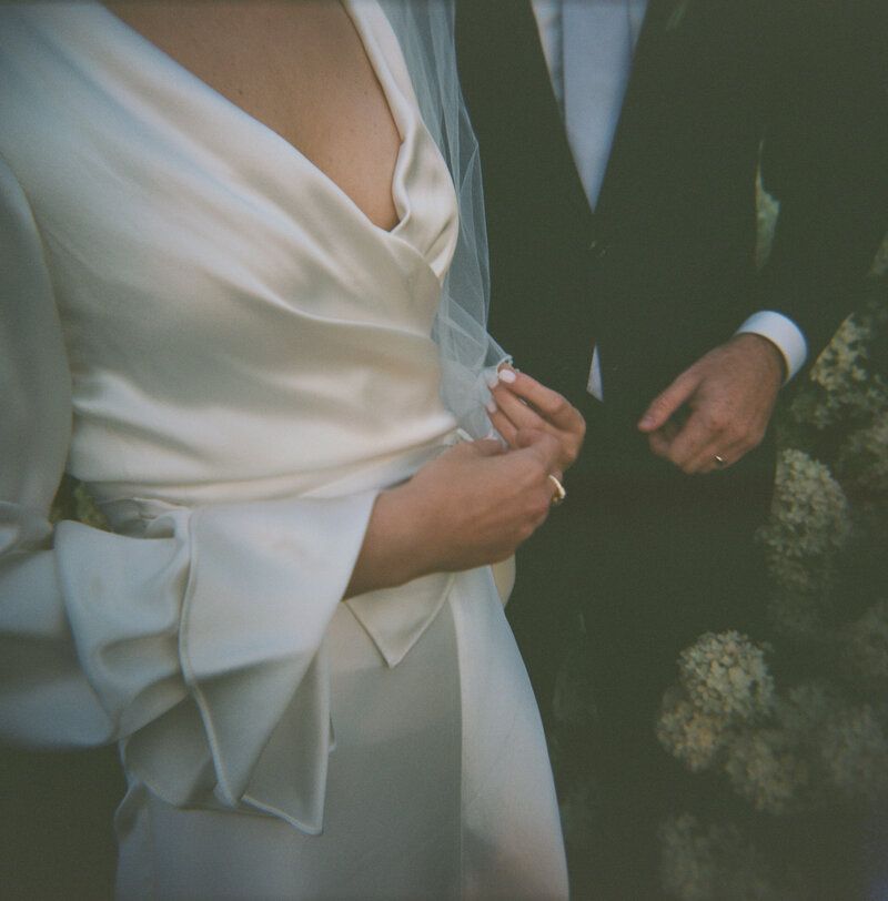 Bride in elegant white satin wrap wedding dress with bow detail stands beside groom in dark suit holding flowers.