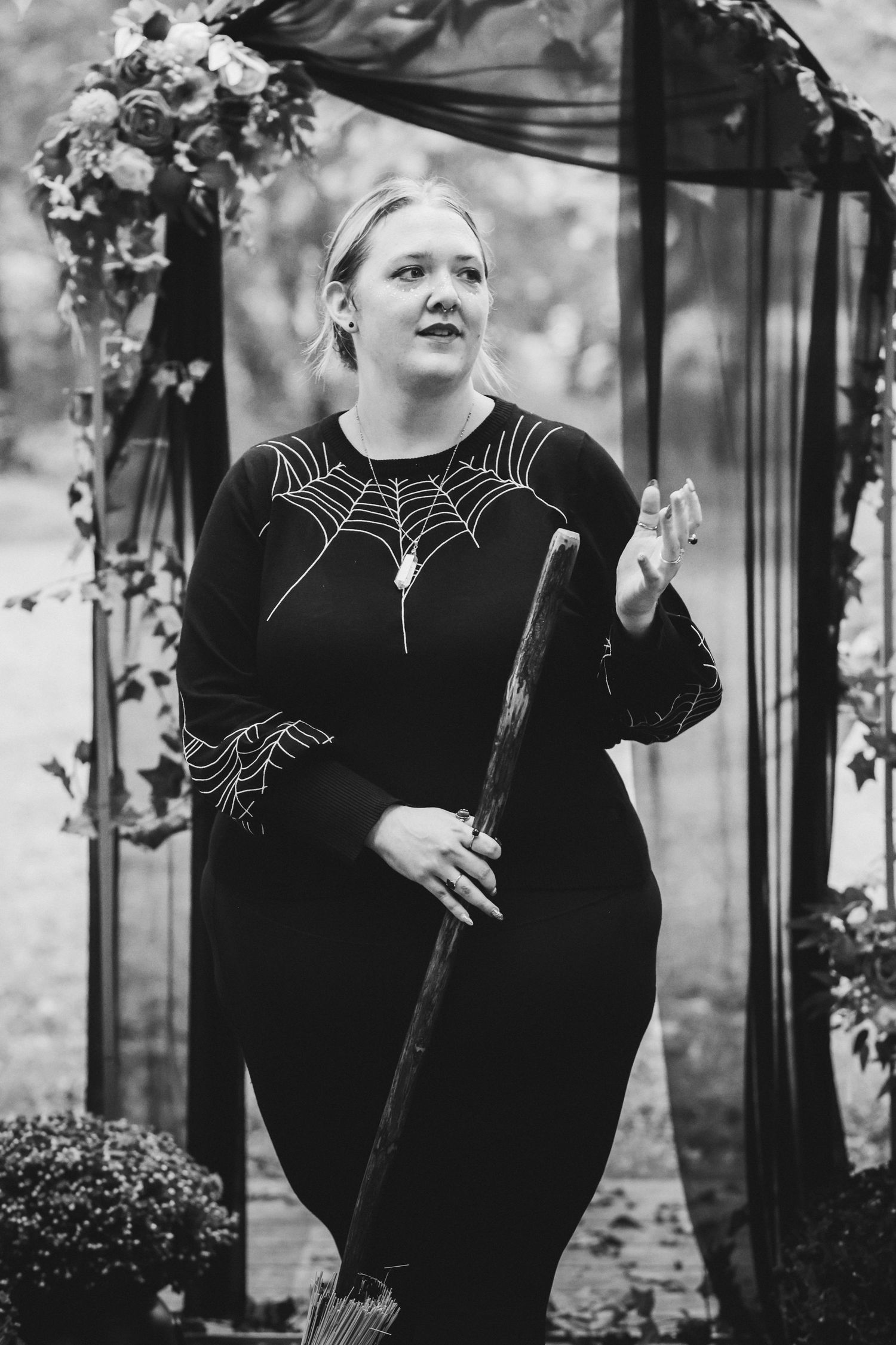Woman in black spider web dress holding a wooden staff stands under a floral arch in a gothic outdoor setting.