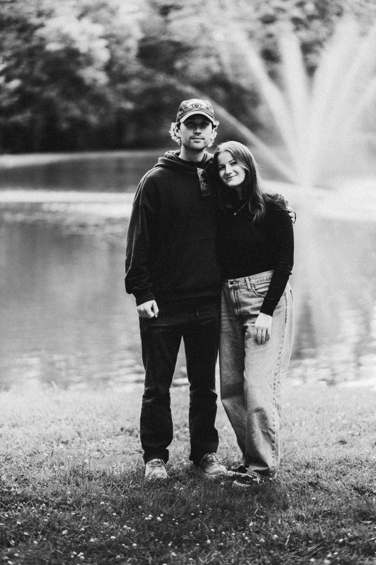 Black and white photo of a couple standing together by a pond with a fountain, smiling at the camera outdoors.