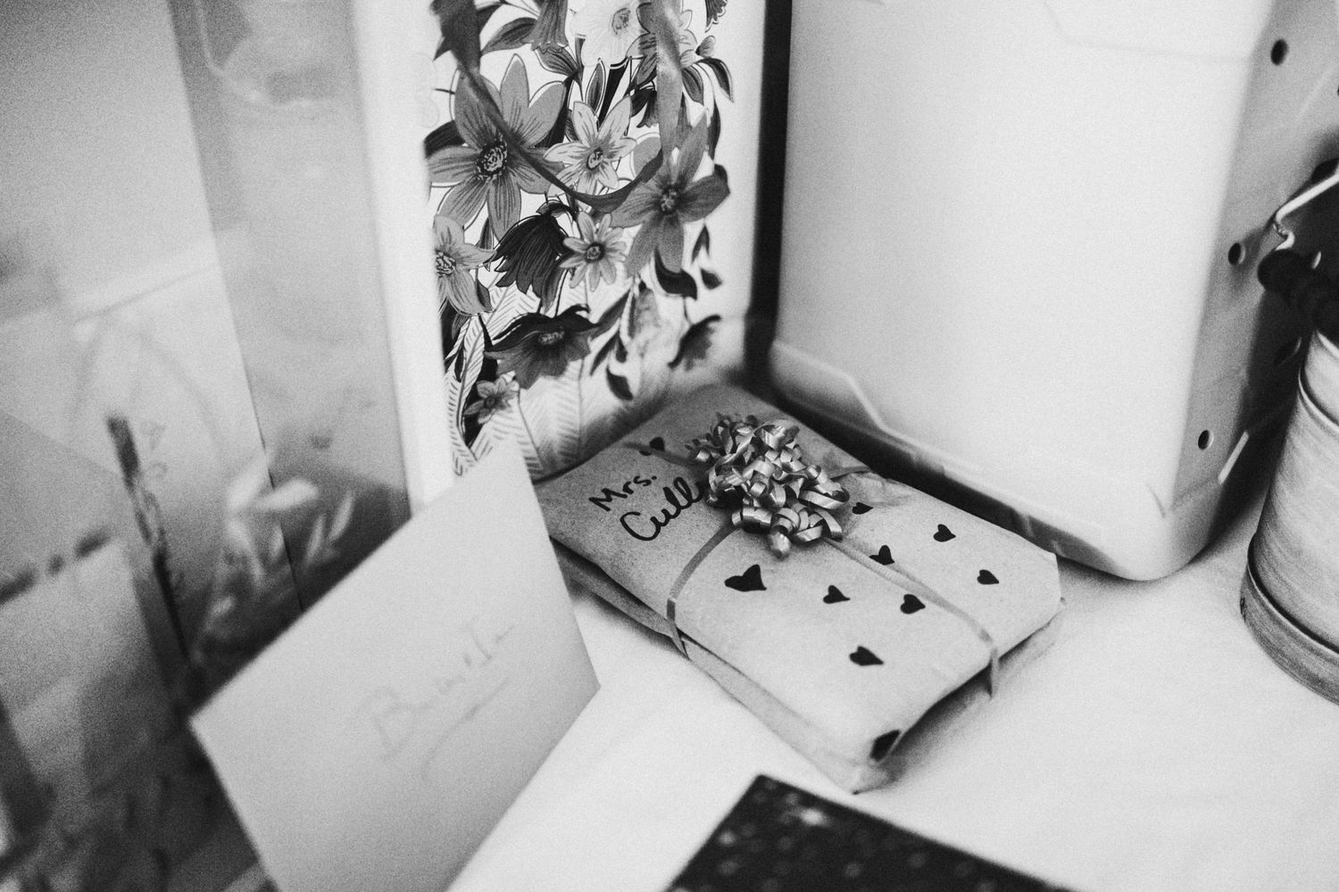 Black and white photo of wrapped gifts with bow, floral artwork, and handwritten note on a shelf.