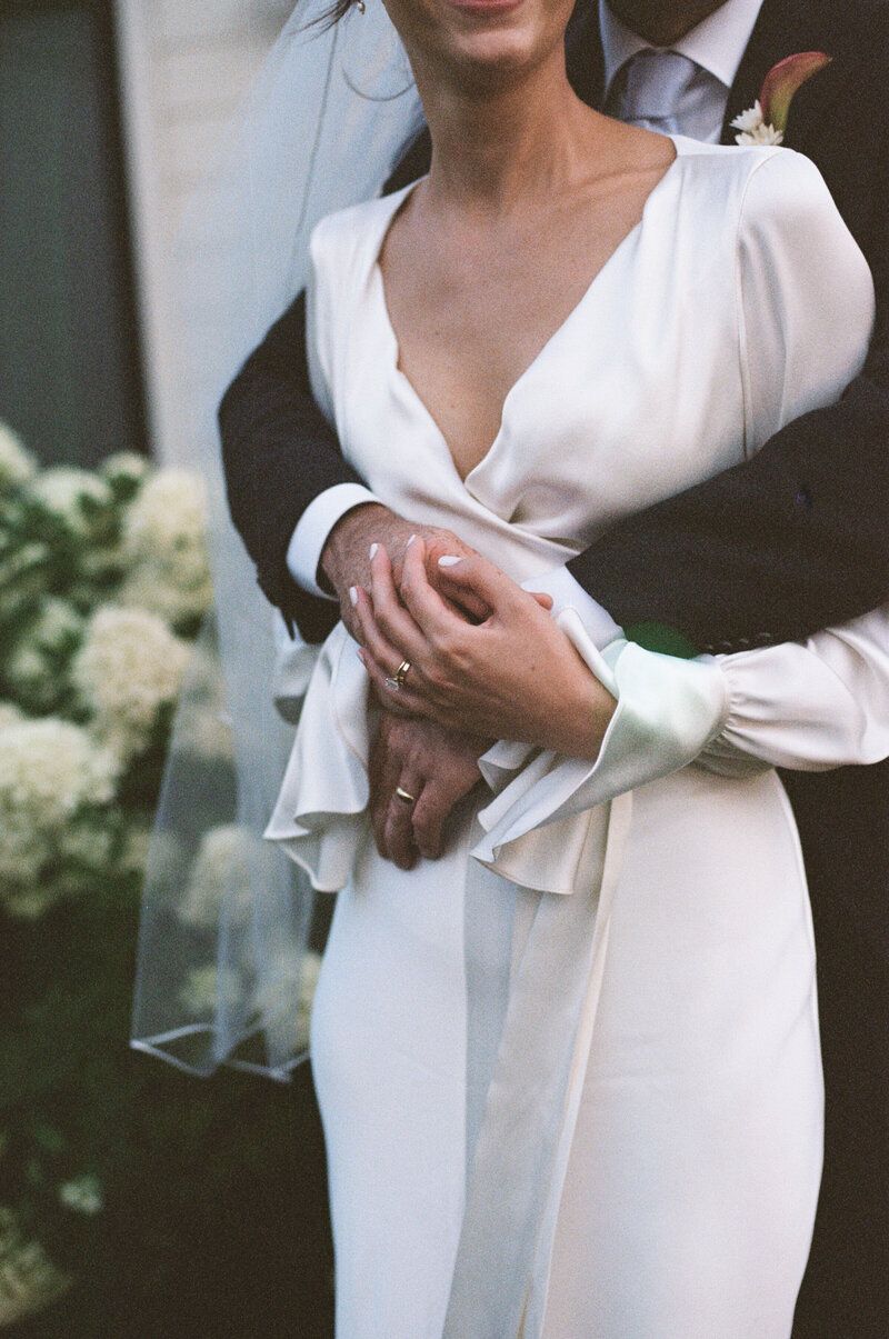 Bride in white wrap wedding dress with veil held by groom in suit, both wearing wedding rings, near white flowers.