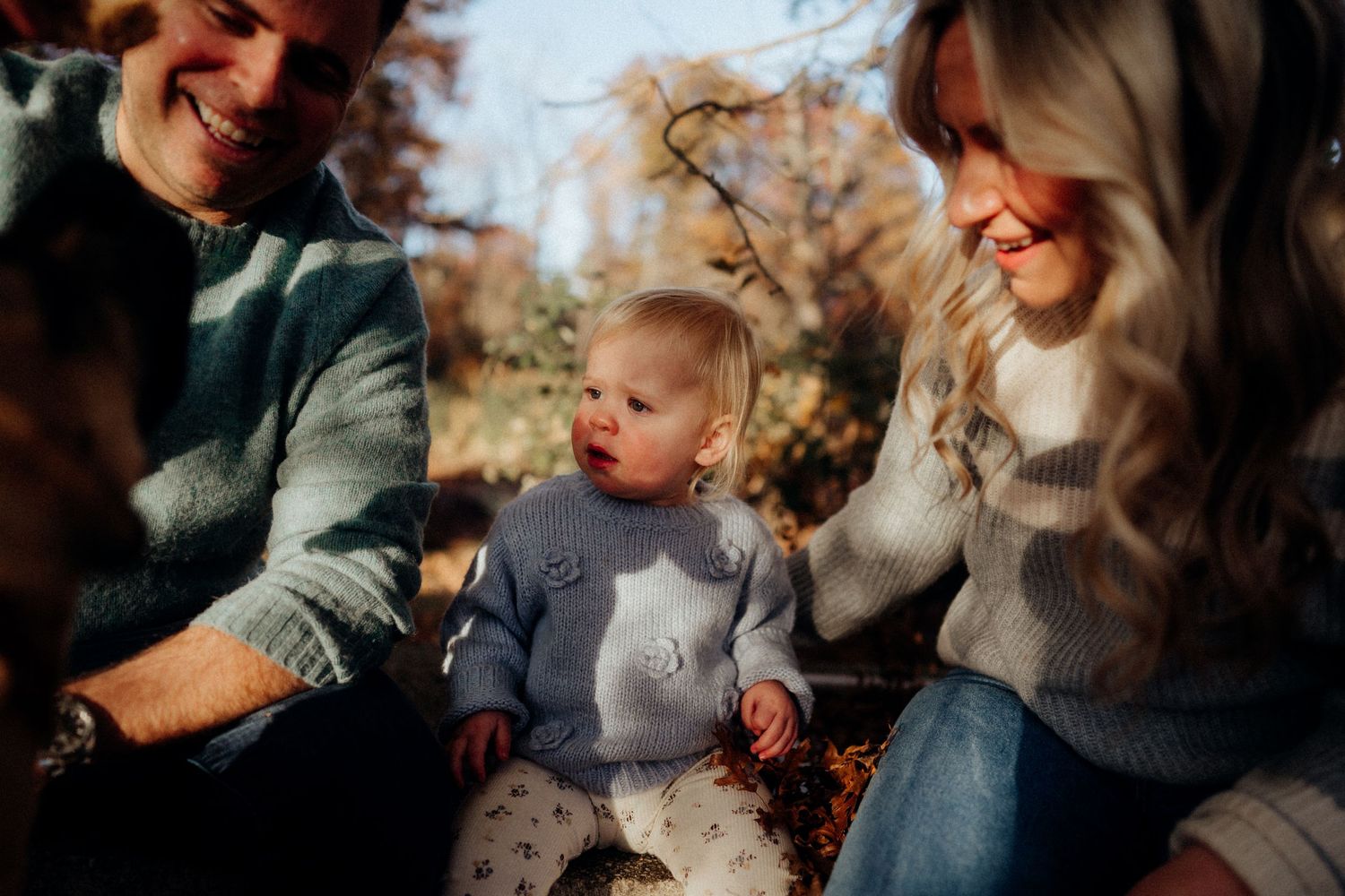 A young child in a gray sweater sits between adults outdoors during autumn.