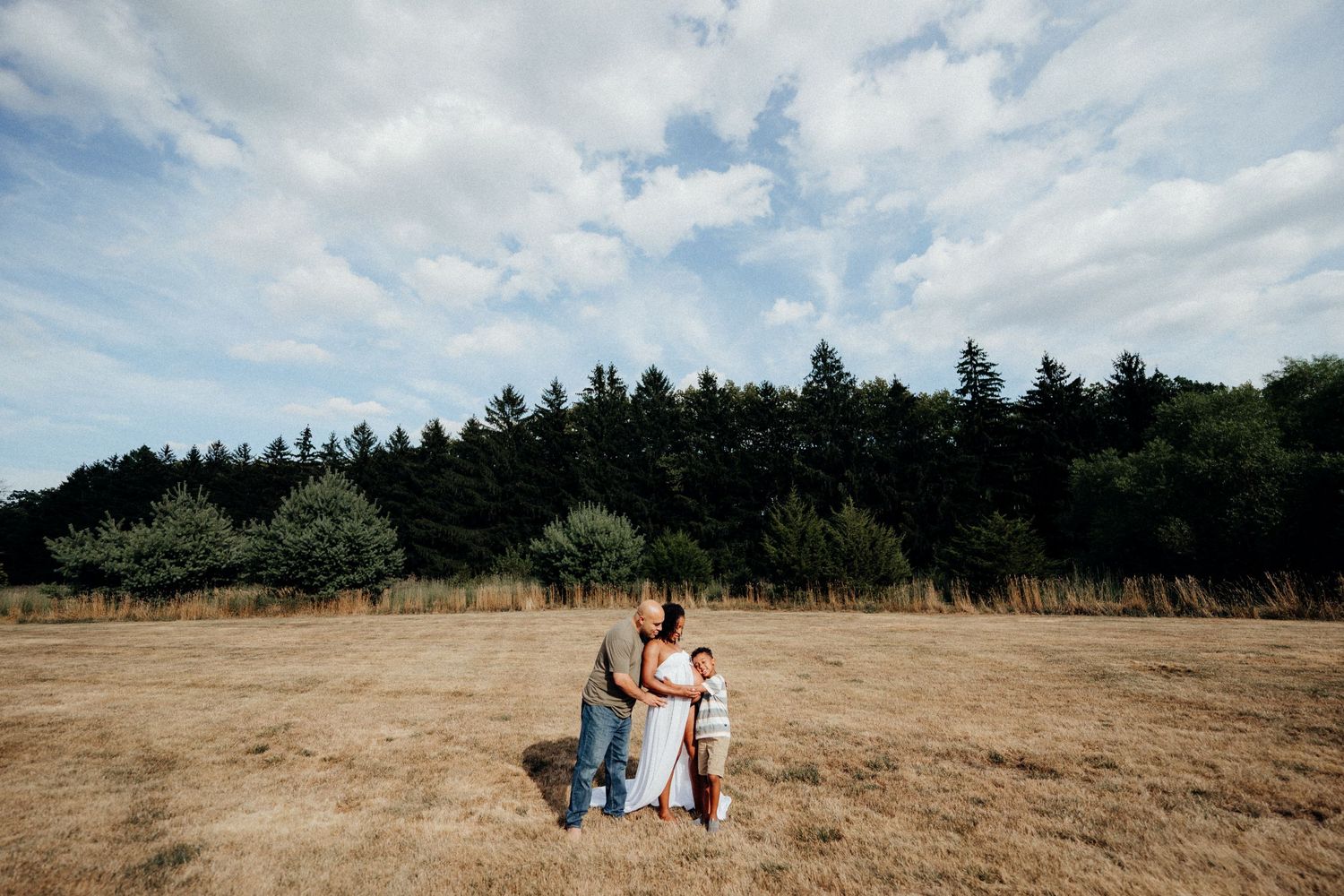 Two people stand together in an open field with trees and blue sky in the background.