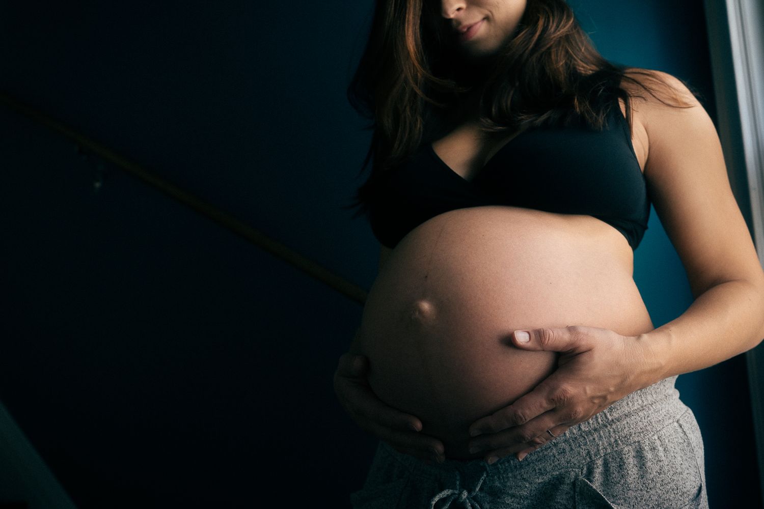A pregnant woman in black top and gray pants gently cradles her baby bump in moody lighting.