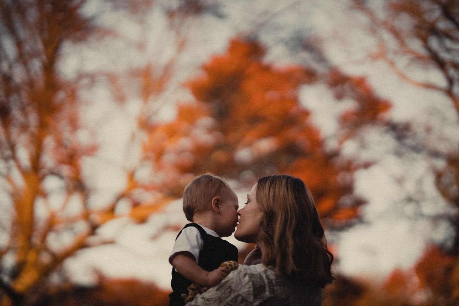 Silhouetted figures share a tender moment against a backdrop of autumn trees with orange foliage at sunset.