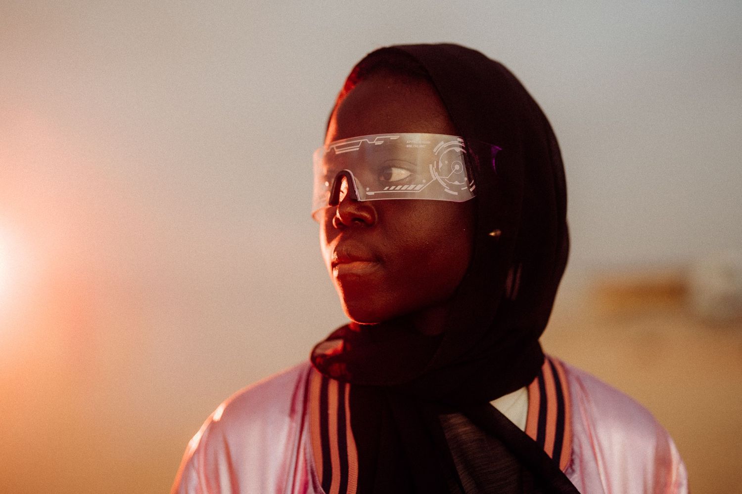 Person wearing futuristic glasses and hijab illuminated by warm side lighting.