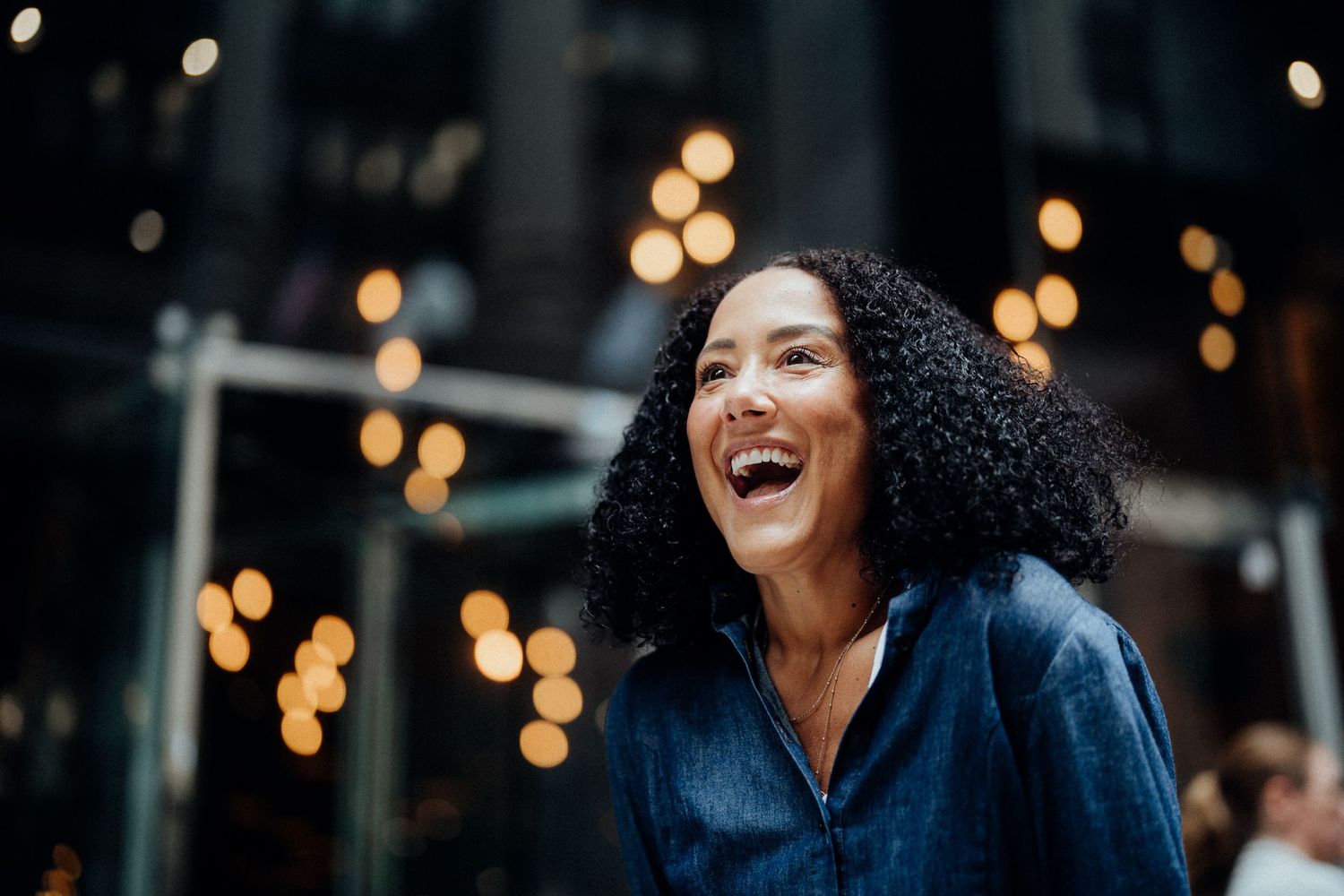 Person with curly hair laughing joyfully against evening city lights.