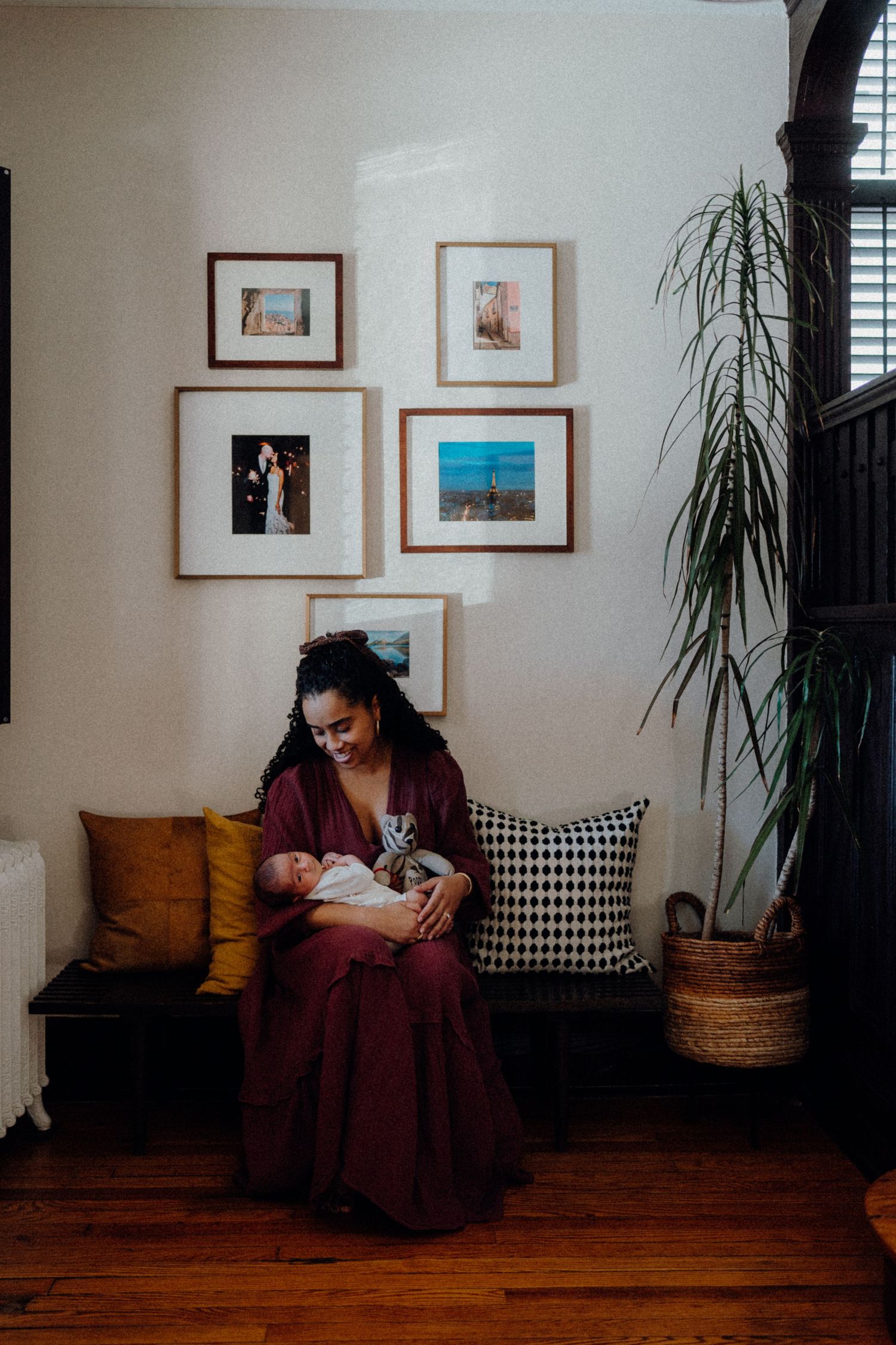 A nursing mother in burgundy dress sits beneath gallery wall with indoor plant.