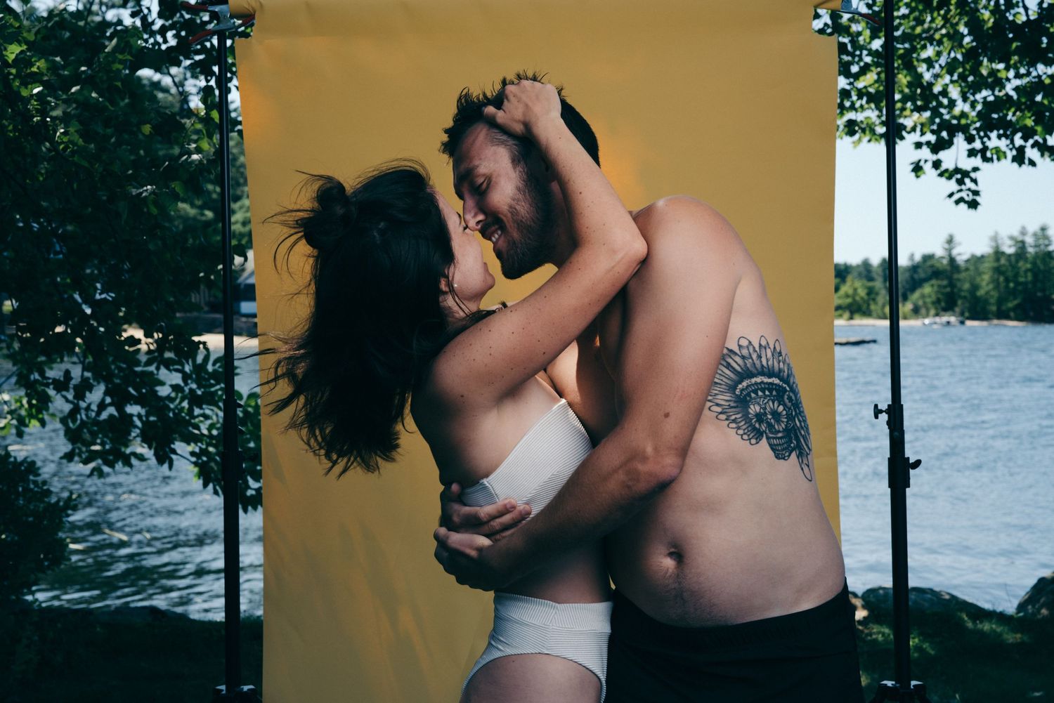 A couple embraces intimately against a yellow backdrop near a waterfront setting.