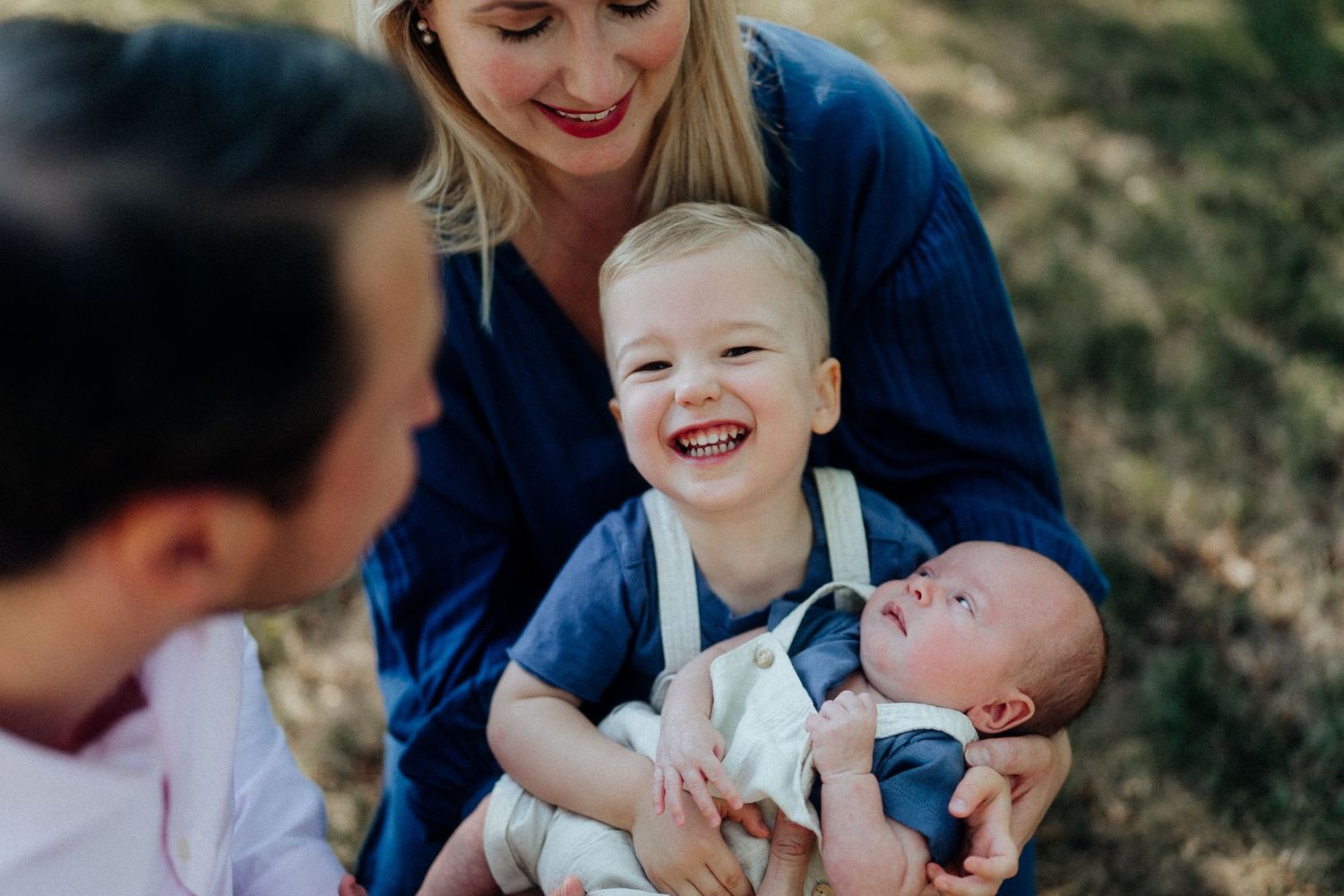 Family sitting outdoors on grass with laughing toddler and newborn baby.