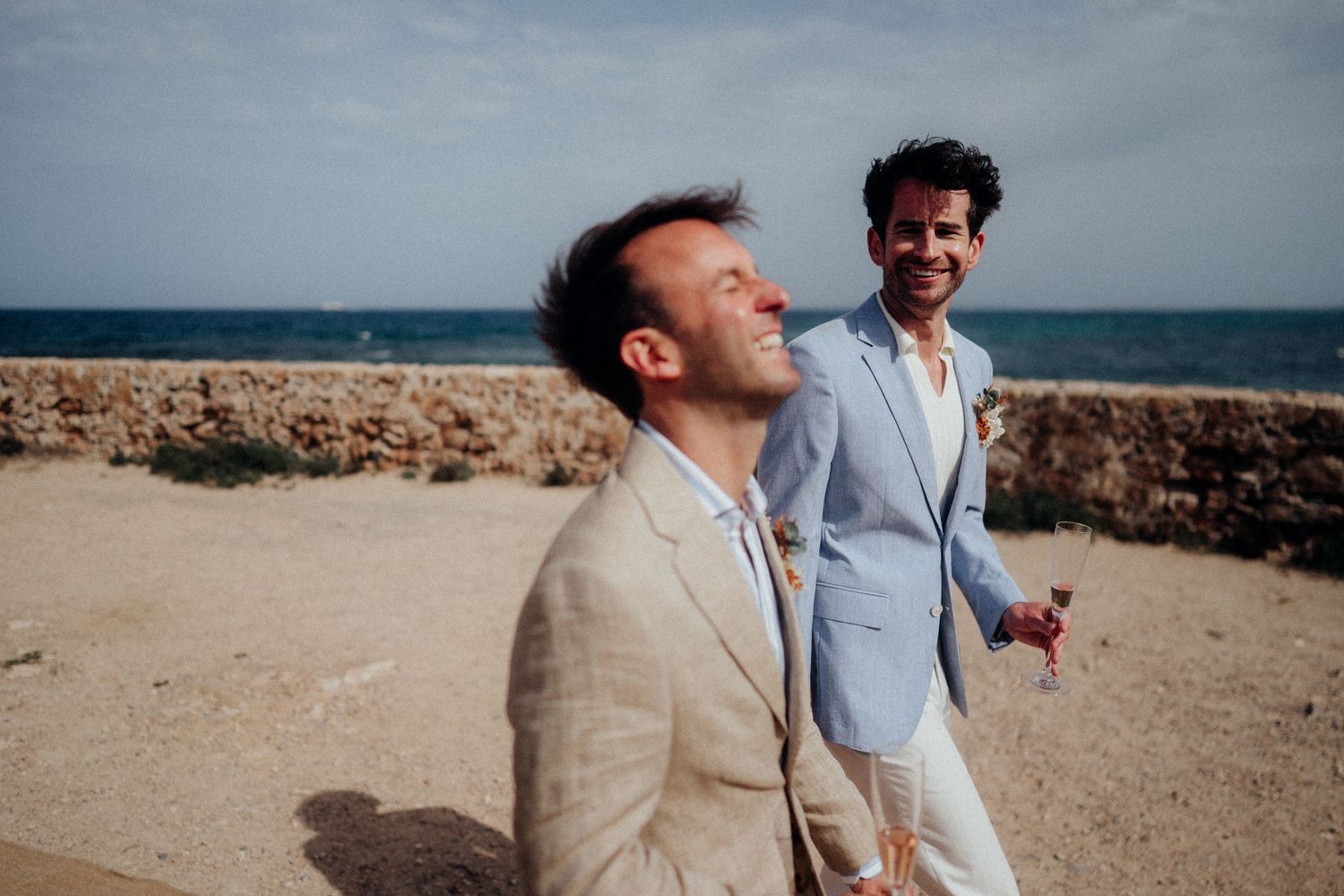 Two people in light suits share a candid moment by the ocean.
