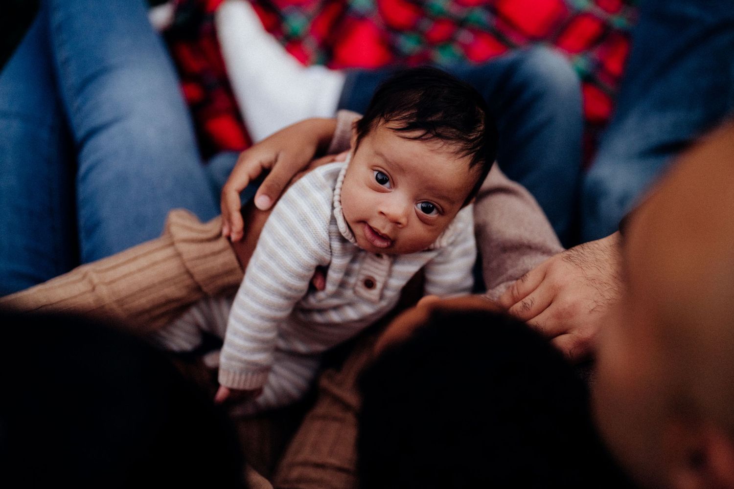 A small baby in a striped sweater being held by adult hands against a red plaid background.