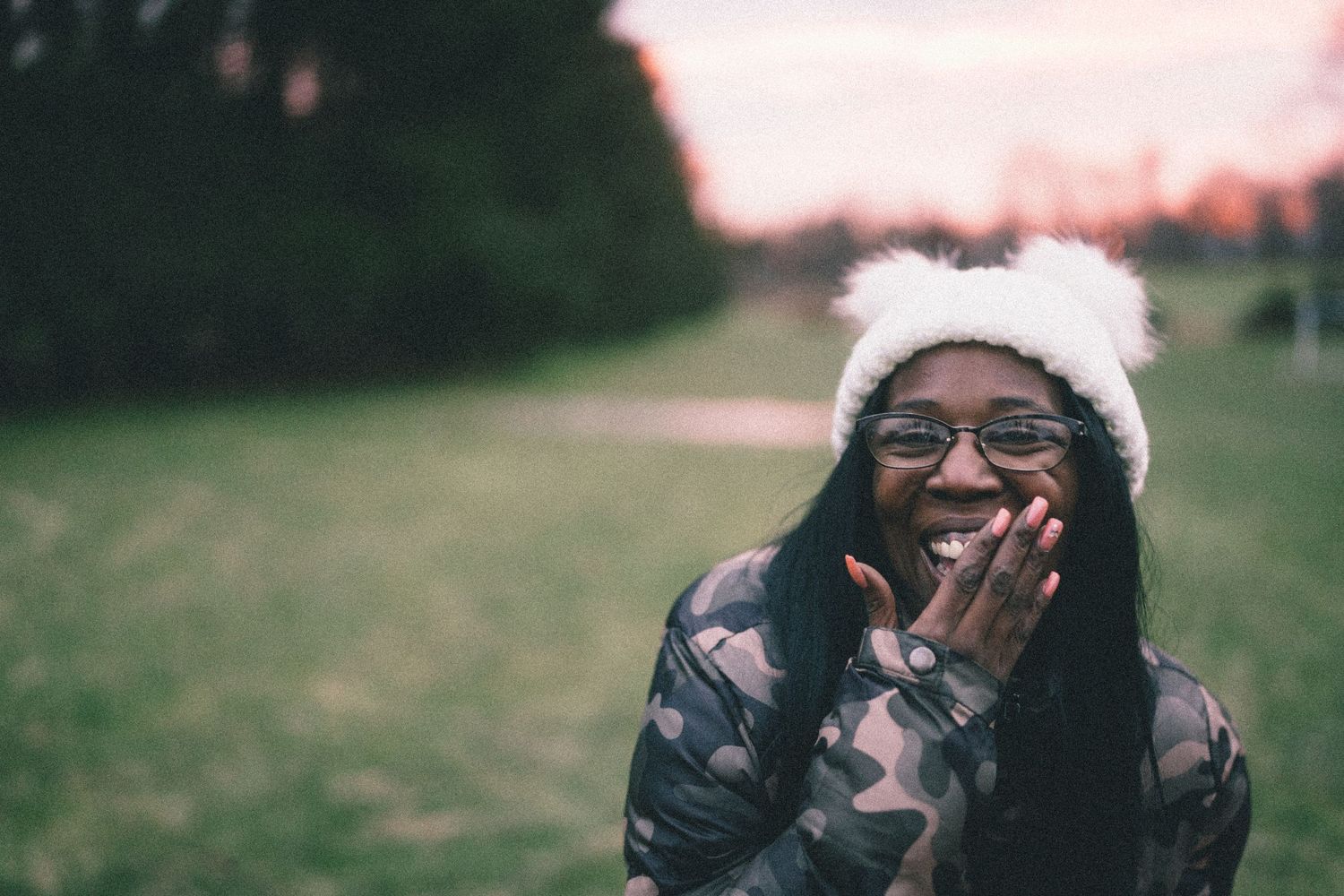 Person wearing a fuzzy white hat and glasses laughing outdoors at sunset.
