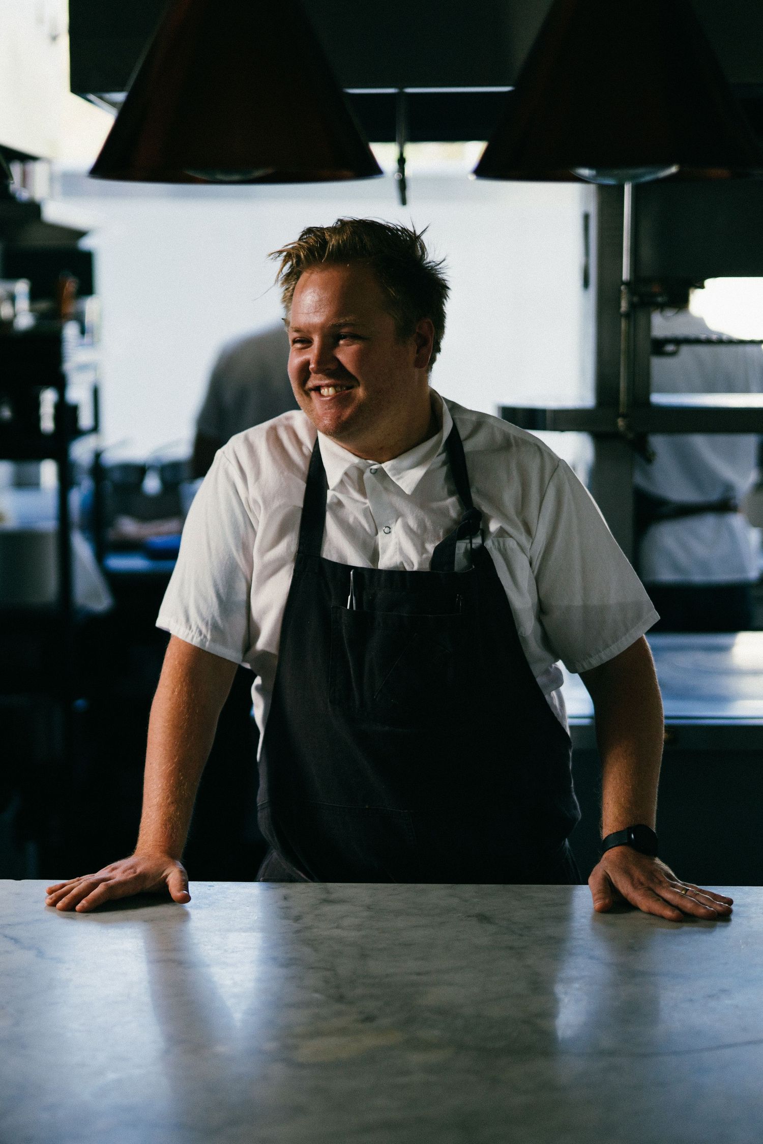 Chef wearing black apron leans on metal counter with warm smile in restaurant kitchen.