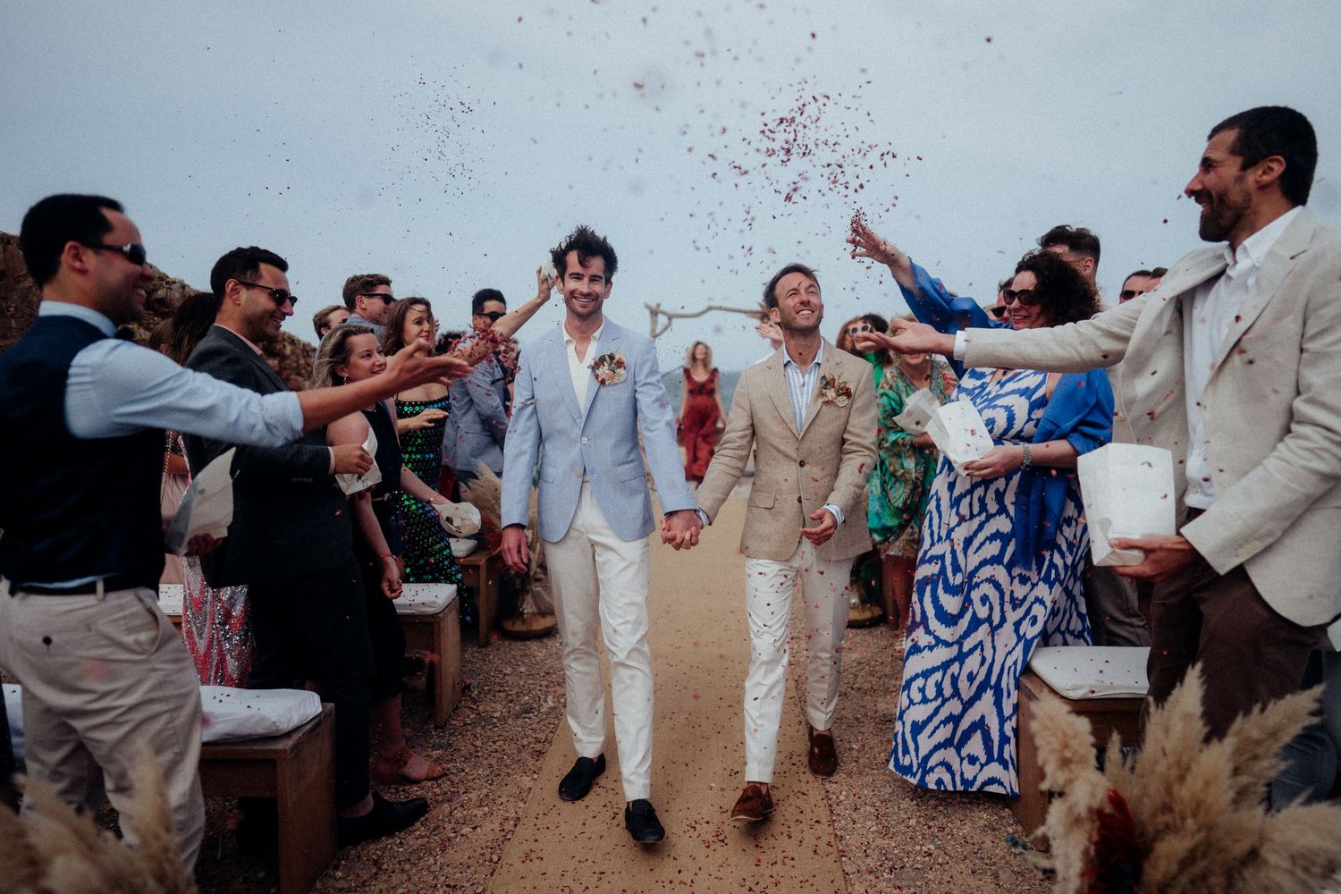 Wedding guests throw confetti over celebrating newlyweds walking down an outdoor aisle.