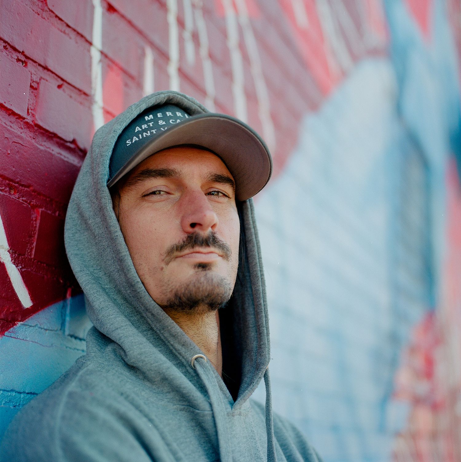 Person in gray hoodie and baseball cap leaning against red wall with urban backdrop.