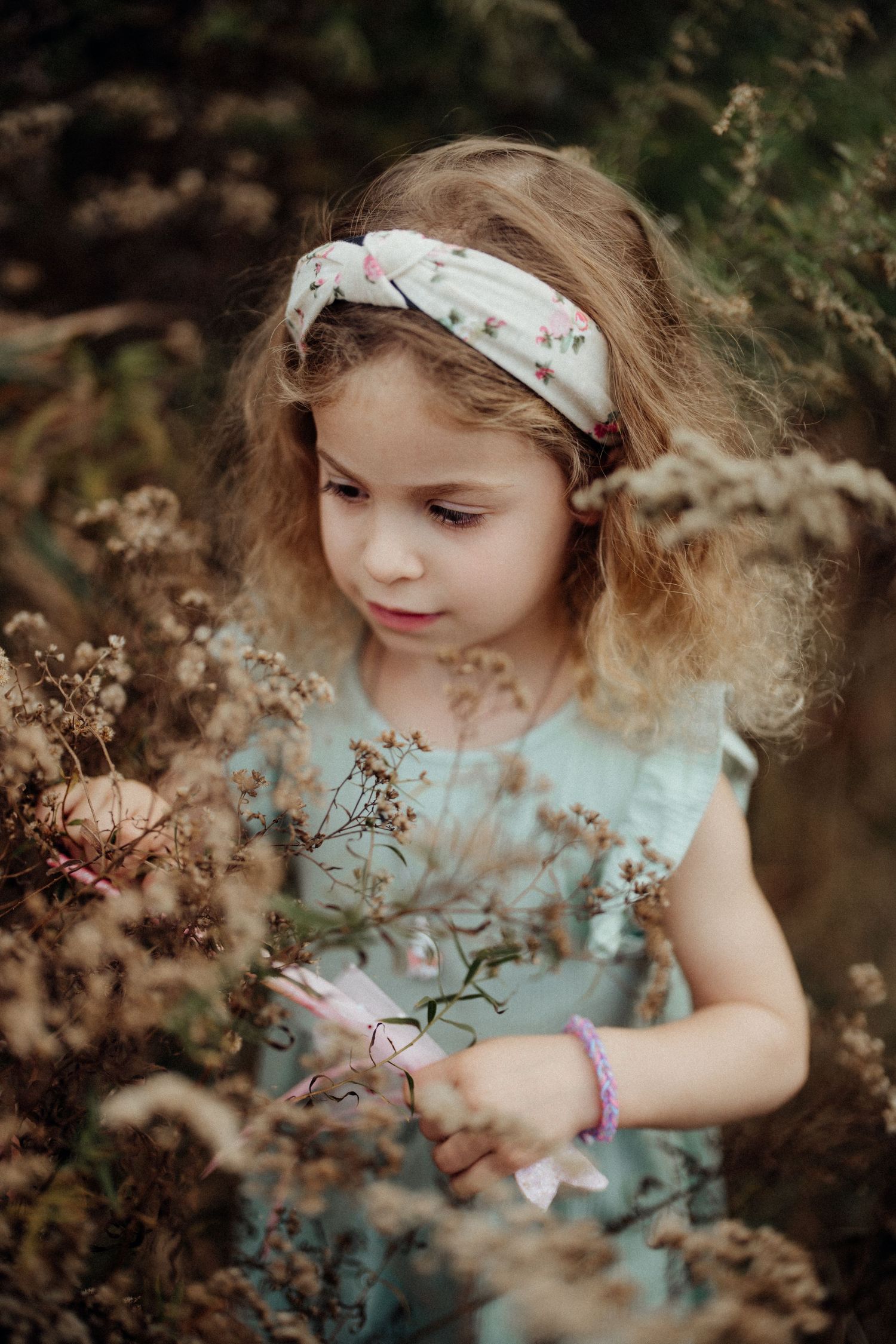 A young child in a floral dress and headband explores wildflowers in a garden setting.