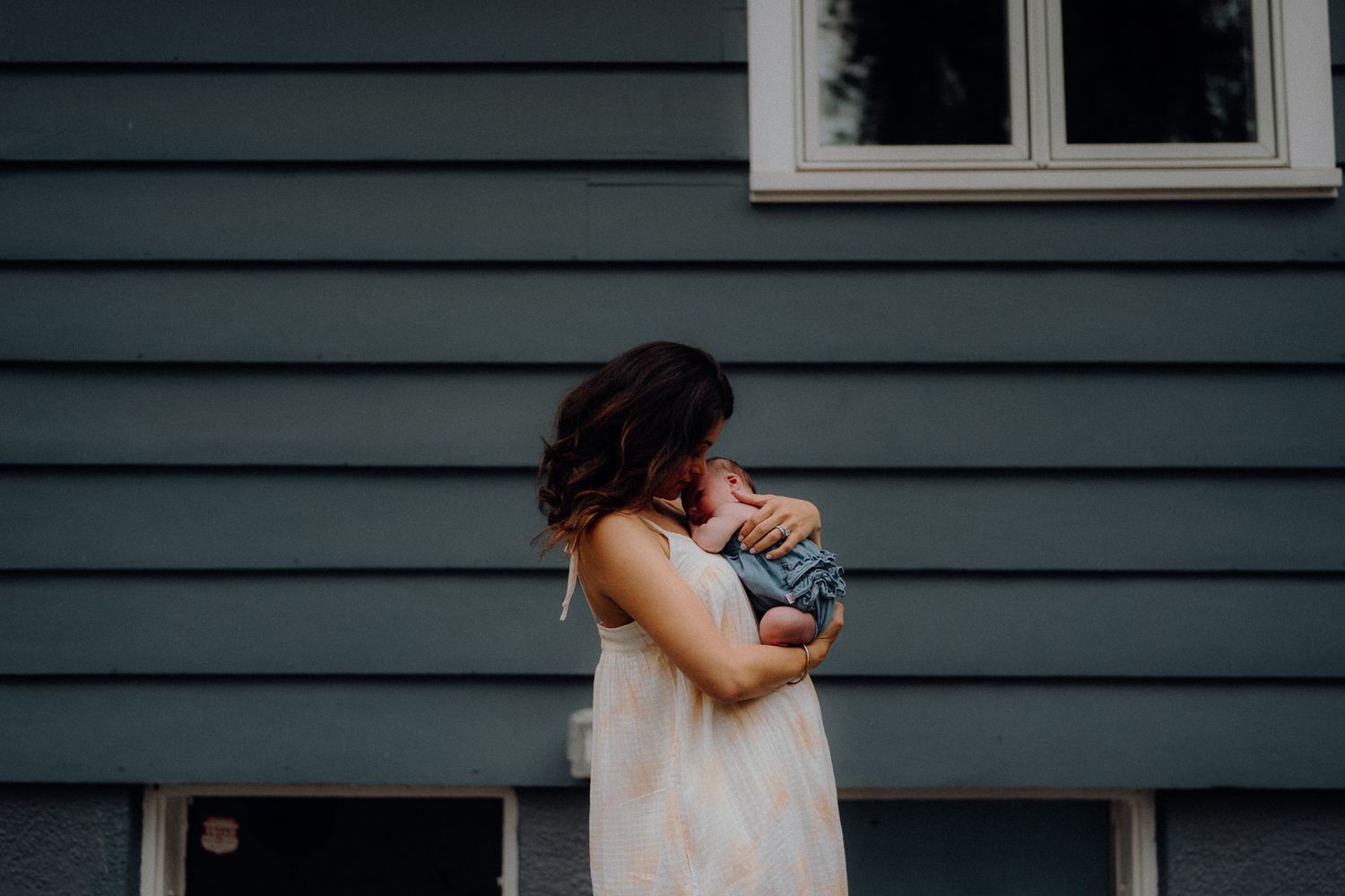 A figure in a white dress holds a small baby against a blue house exterior.