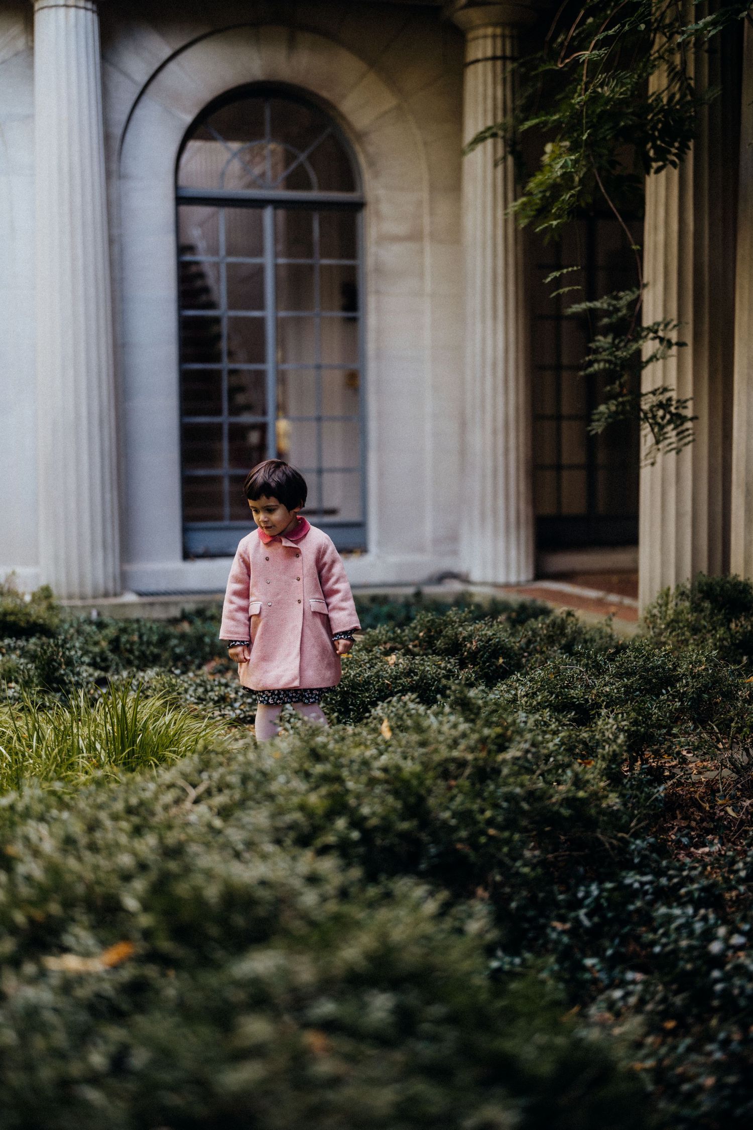 A small child in a pink coat standing among bushes near a classical building with columns.