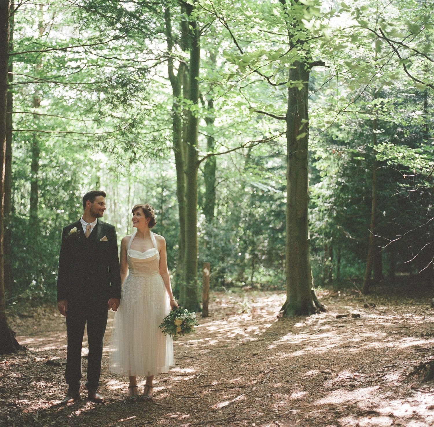 Newlywed couple walks through sunlit forest in romantic wedding attire.
