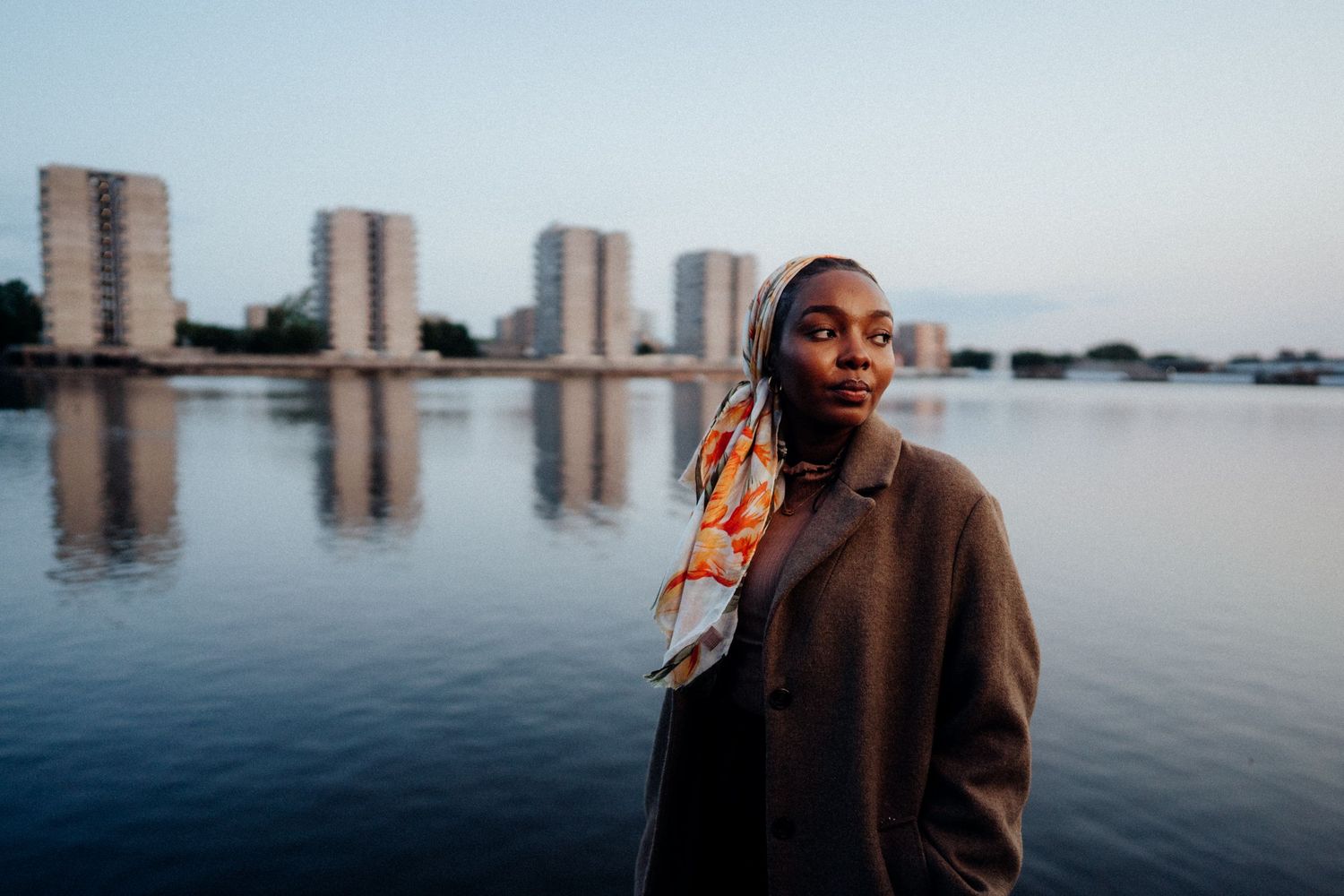 Person in brown coat standing by waterfront with city buildings reflected in calm water.