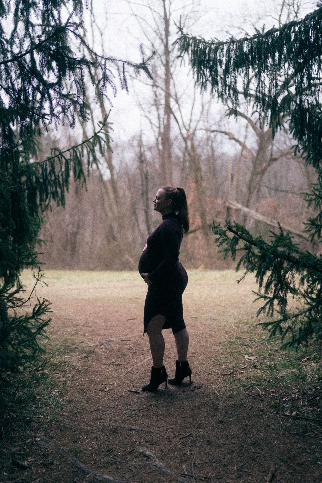 Silhouette of an expectant mother standing between evergreen trees in a misty forest.