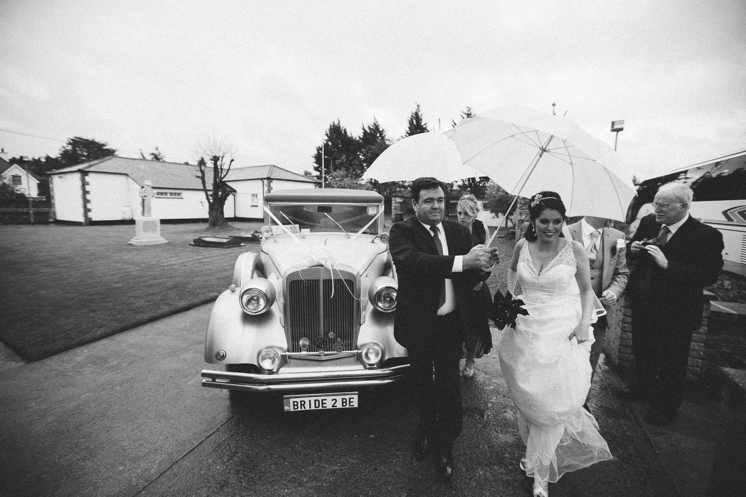 A classic white wedding car with guests holding umbrellas on a rainy day.