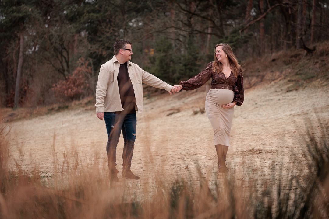 zwangerschapsfotoshoot waarbij partners elkaar speels aankijken en over het strand van de Bergse Heide lopen
