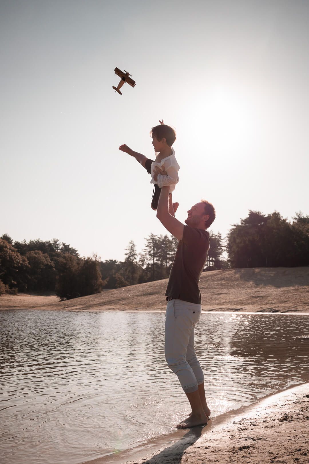 Speelmoment tijdens gezinsfotoshoot aan het water, vader gooit zijn zoontje in de lucht, zoontje gooit speelgoed vliegtuigje in de lucht