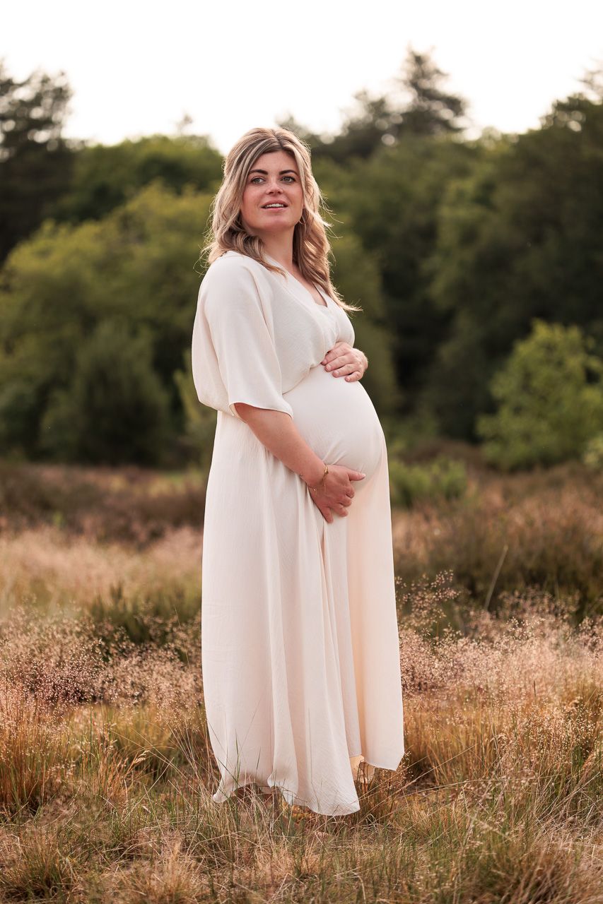 zwangere vrouw in zwierige jurk tussen het gras met haar handen om haar zwangere buik door fotograaf Fotos van Jess