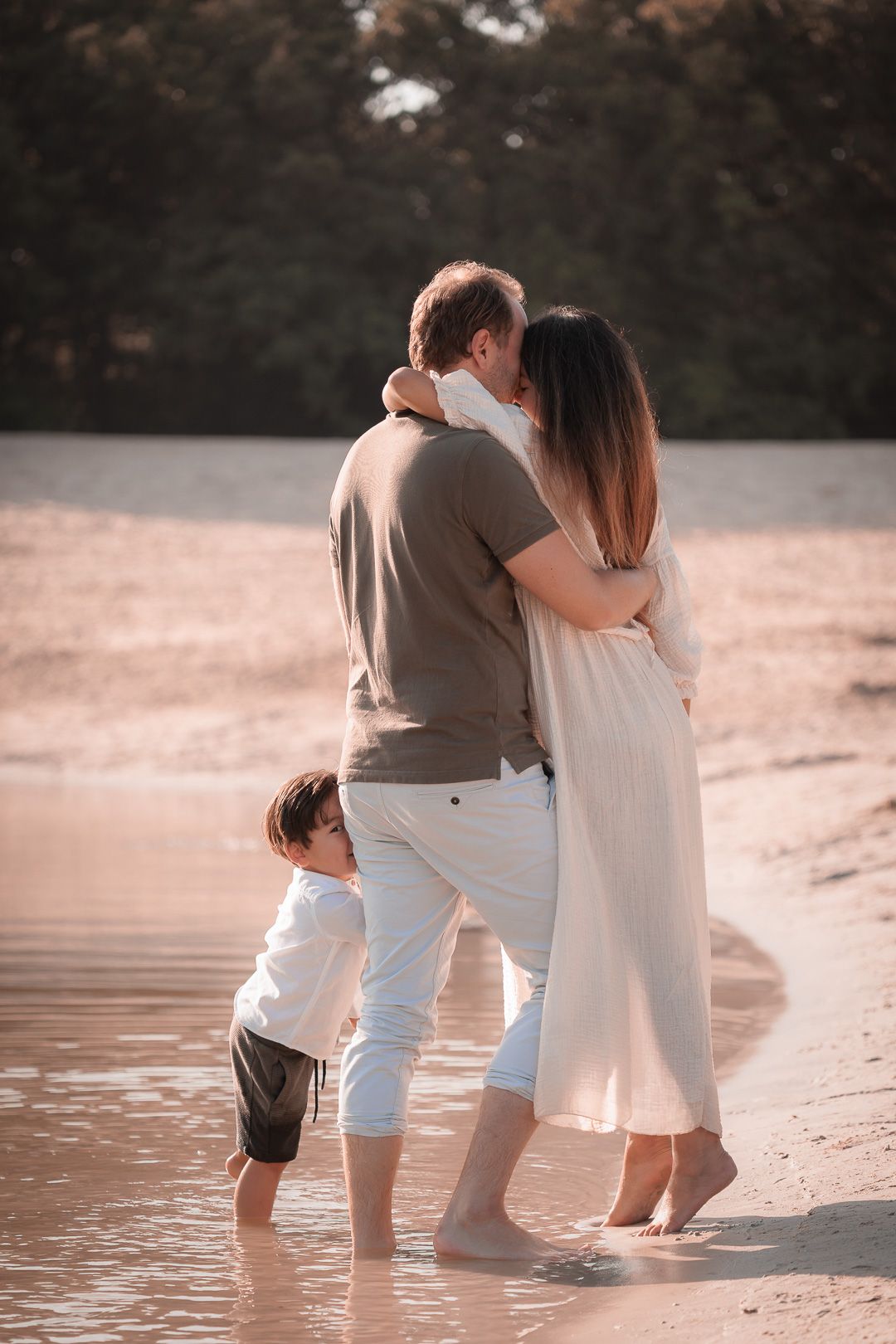 Vader en moeder in omhelzing, zoontje kijkt ondeugend achter zijn vader's been naar de fotograaf tijdens ongedwongen gezinsfotoshoot
