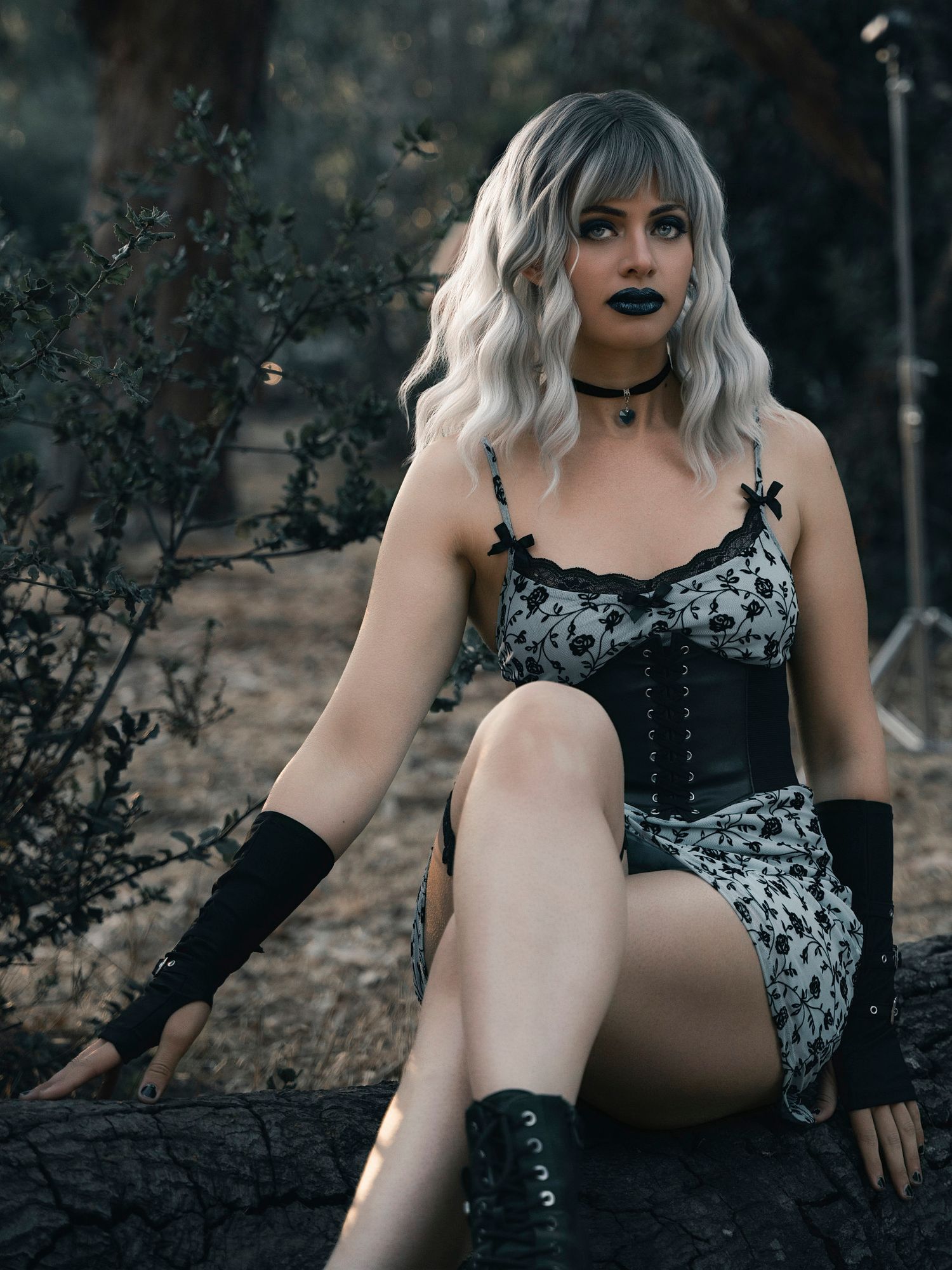 A woman in gothic fashion with platinum hair, black lipstick, corset, and lace dress poses outdoors.